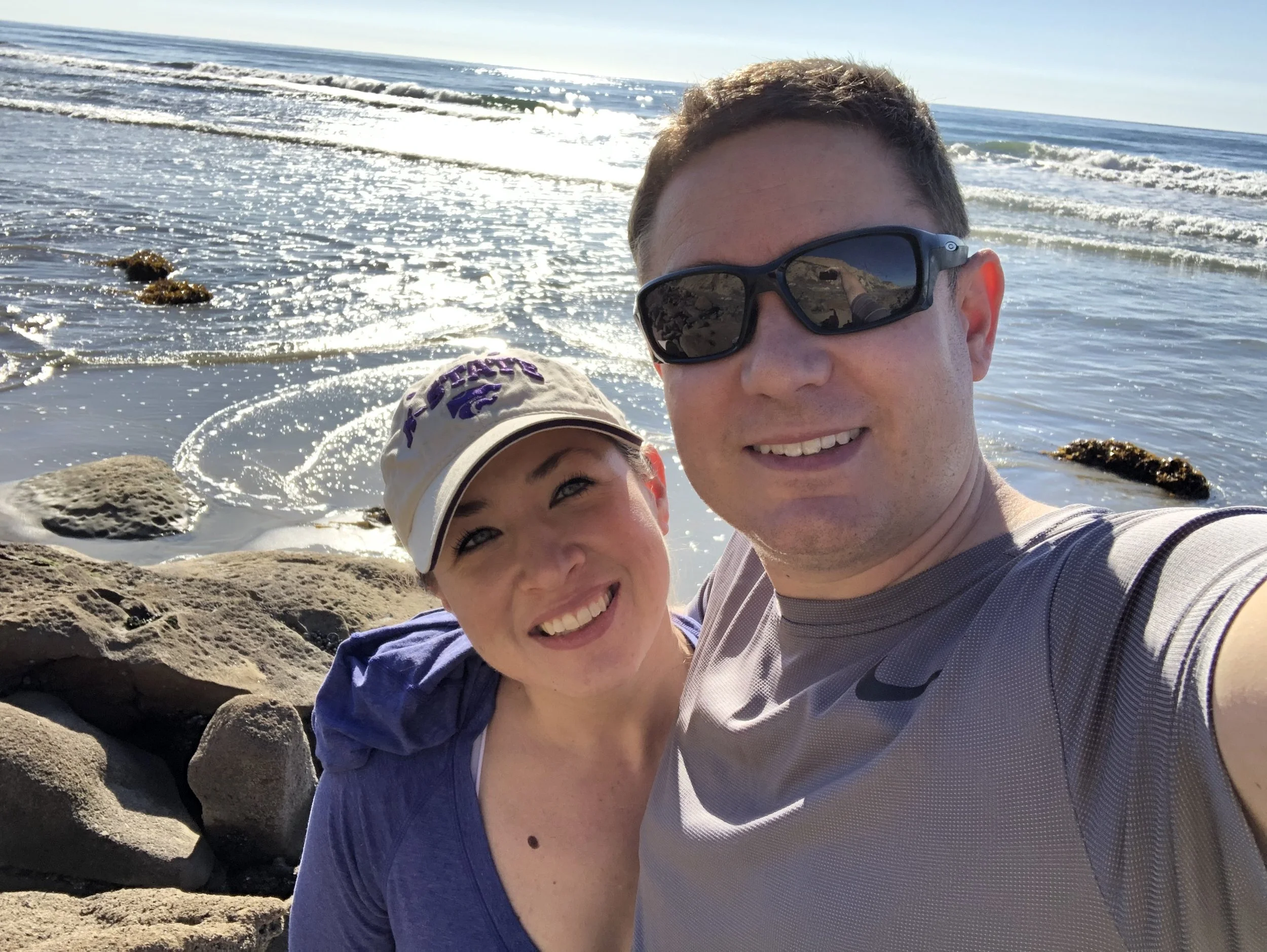 A smiling man and woman taking a selfie on a rocky beach with ocean waves and a clear sky in the background, both wearing sunglasses. considering adoption for her child and learning how to get pregnant help options to create an adoption plan.