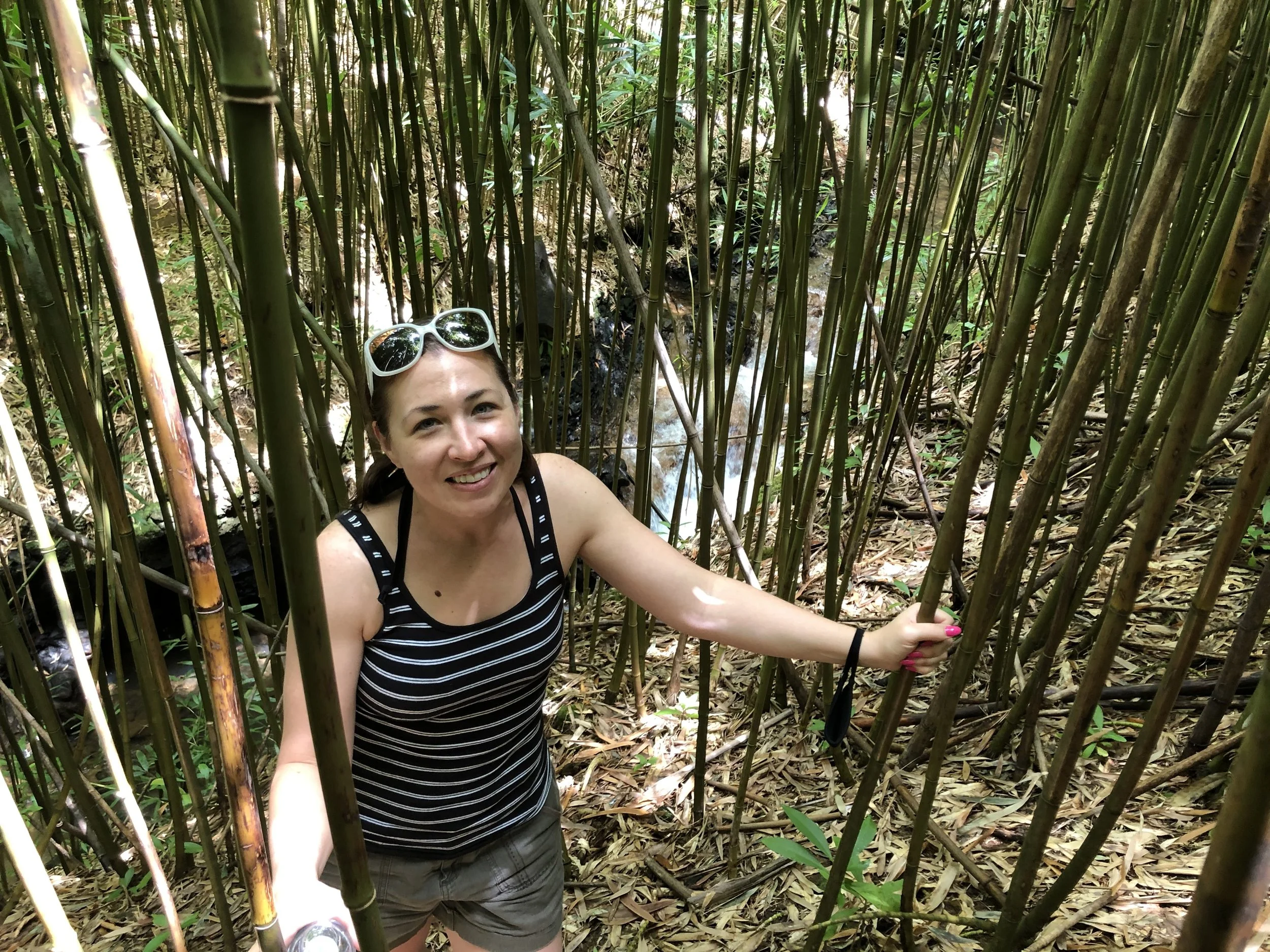A woman with brown hair, wearing sunglasses on her head, a black and white striped tank top, and gray shorts, smiling while standing among dense bamboo stalks in a forest. considering adoption for her child and learning how to get pregnant help optio