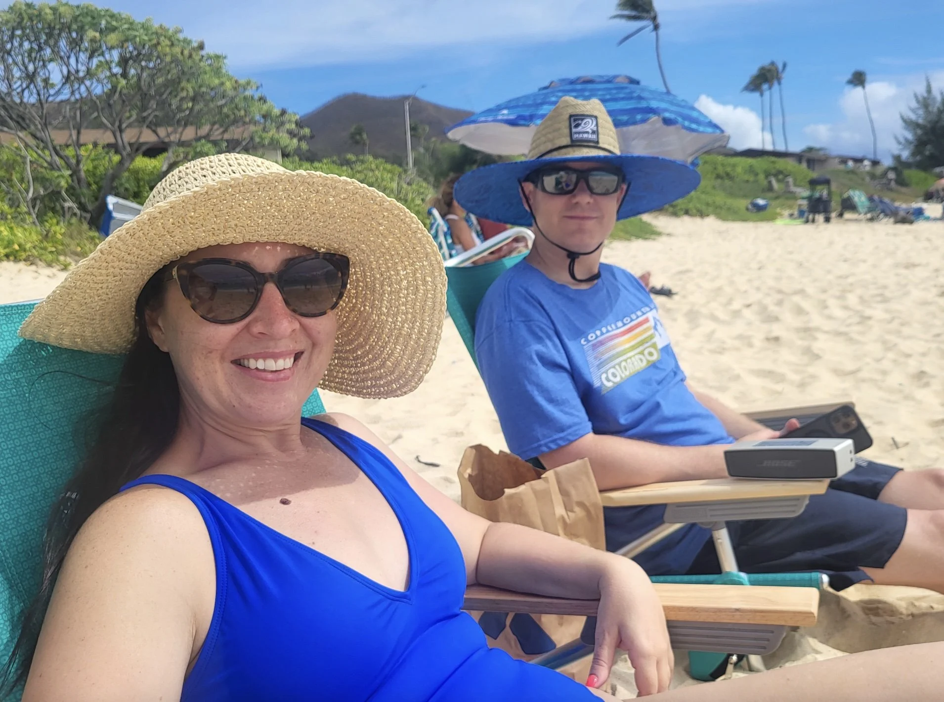 Two people sitting on beach chairs, smiling, wearing large sun hats and sunglasses. The woman in the foreground has long dark hair, is wearing a blue swimsuit, and has a straw sun hat. The man in the background is wearing a blue T-shirt, black sunglasses, and a large blue sun hat with a wide brim and umbrella-like cover. There is sand, greenery, and palm trees in the background, along with a mountain and blue sky.