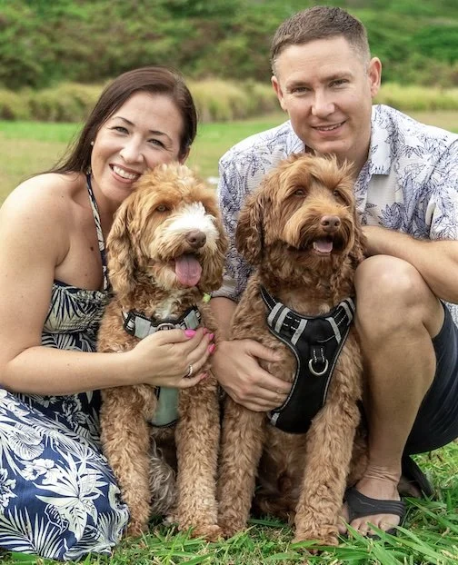 Smiling couple sitting outdoors with two brown Labradoodles in a grassy area. considering adoption for her child and learning how to get pregnant help options to create an adoption plan.