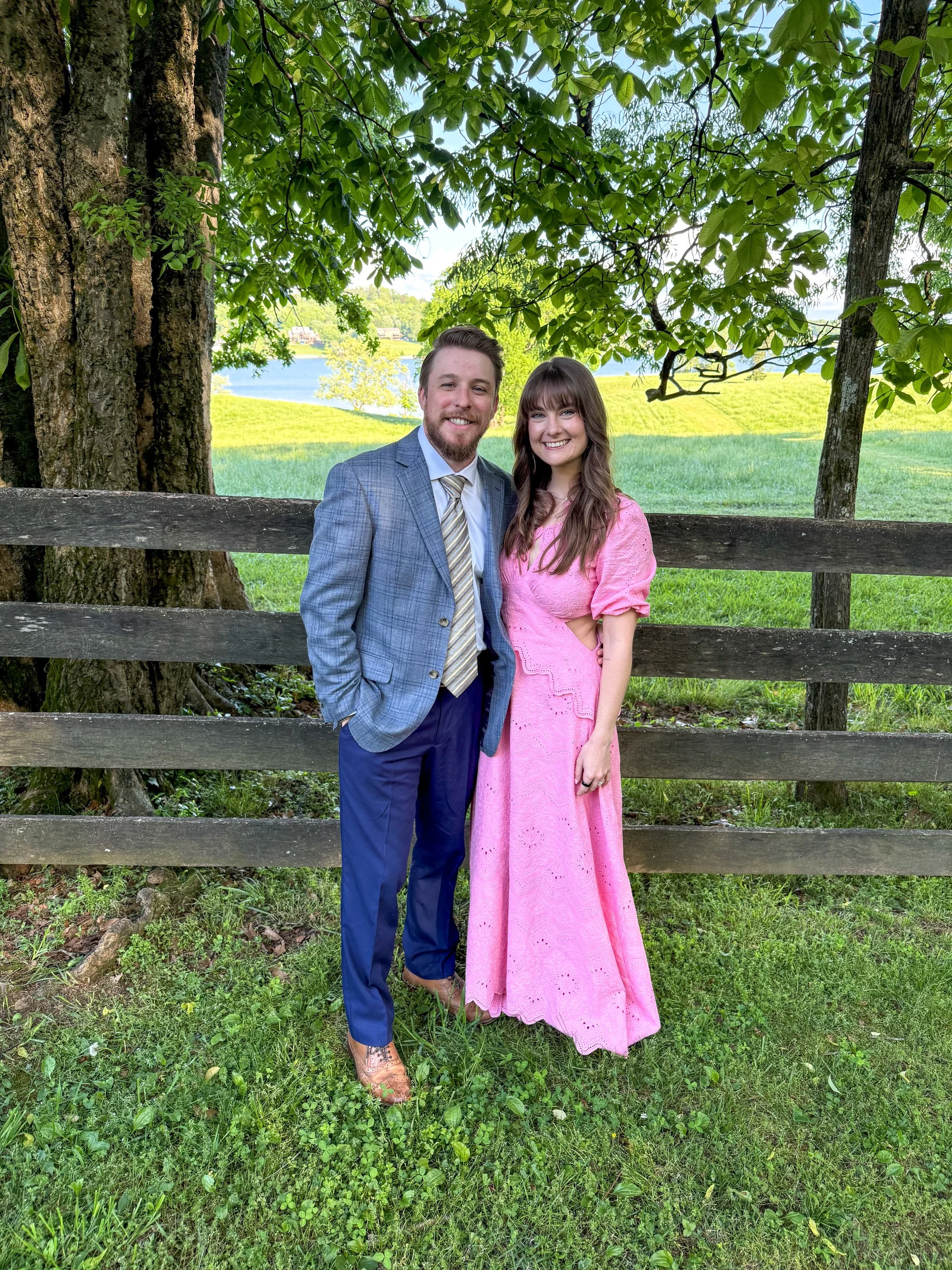 A man and woman standing together outdoors near a wooden fence with greenery, trees, a lake, and a field in the background. The man is wearing a gray suit with a striped tie, and the woman is wearing a pink dress. They are smiling and posing for the photo.