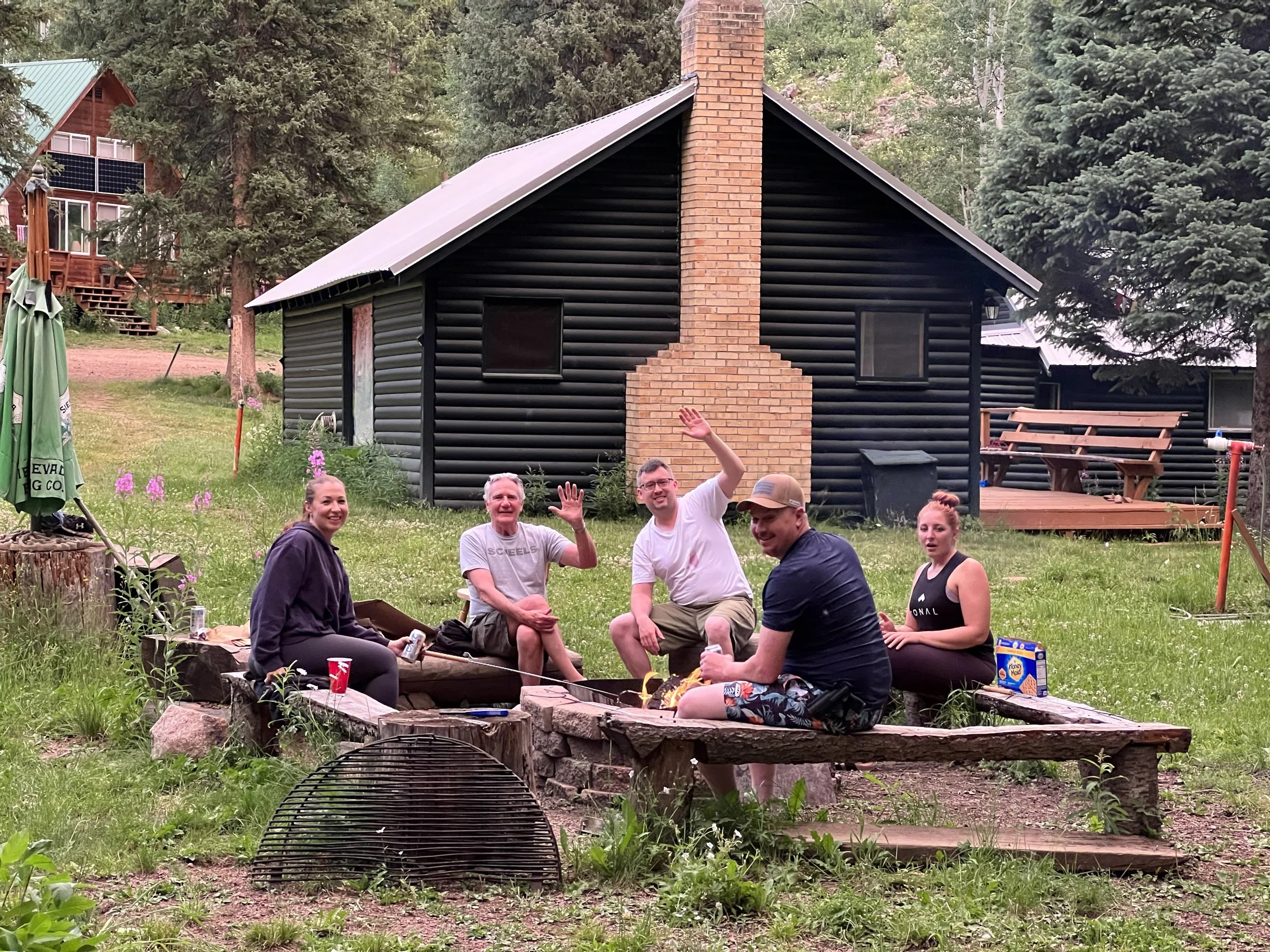 Group of people sitting around a campfire outdoors in a grassy area with forest and cabin in the background, some smiling and waving.