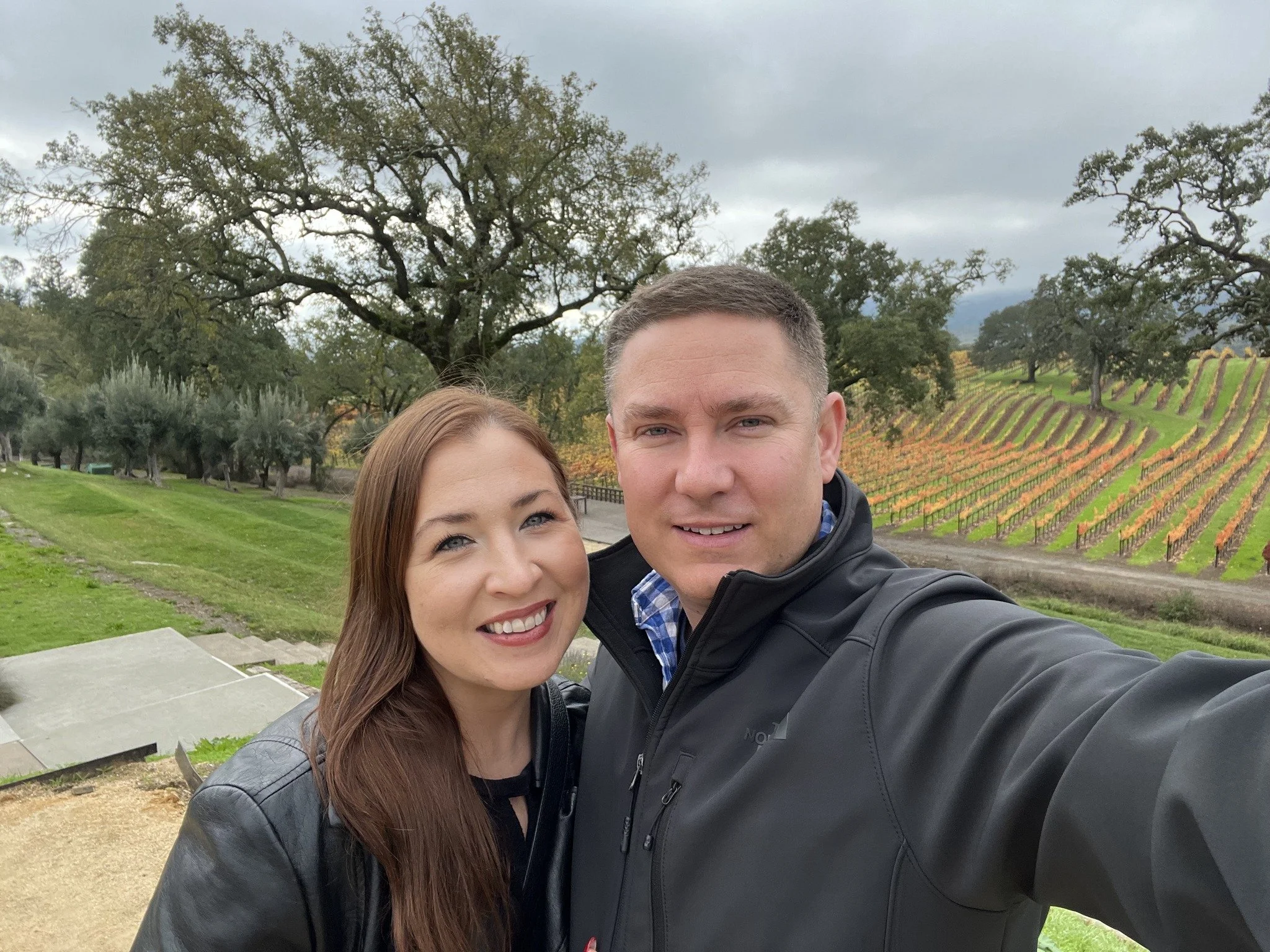 A smiling woman with long brown hair and a smiling man with short hair taking a selfie outdoors in front of a vineyard. There are large trees and a cloudy sky in the background. considering adoption for her child and learning how to get pregnant help