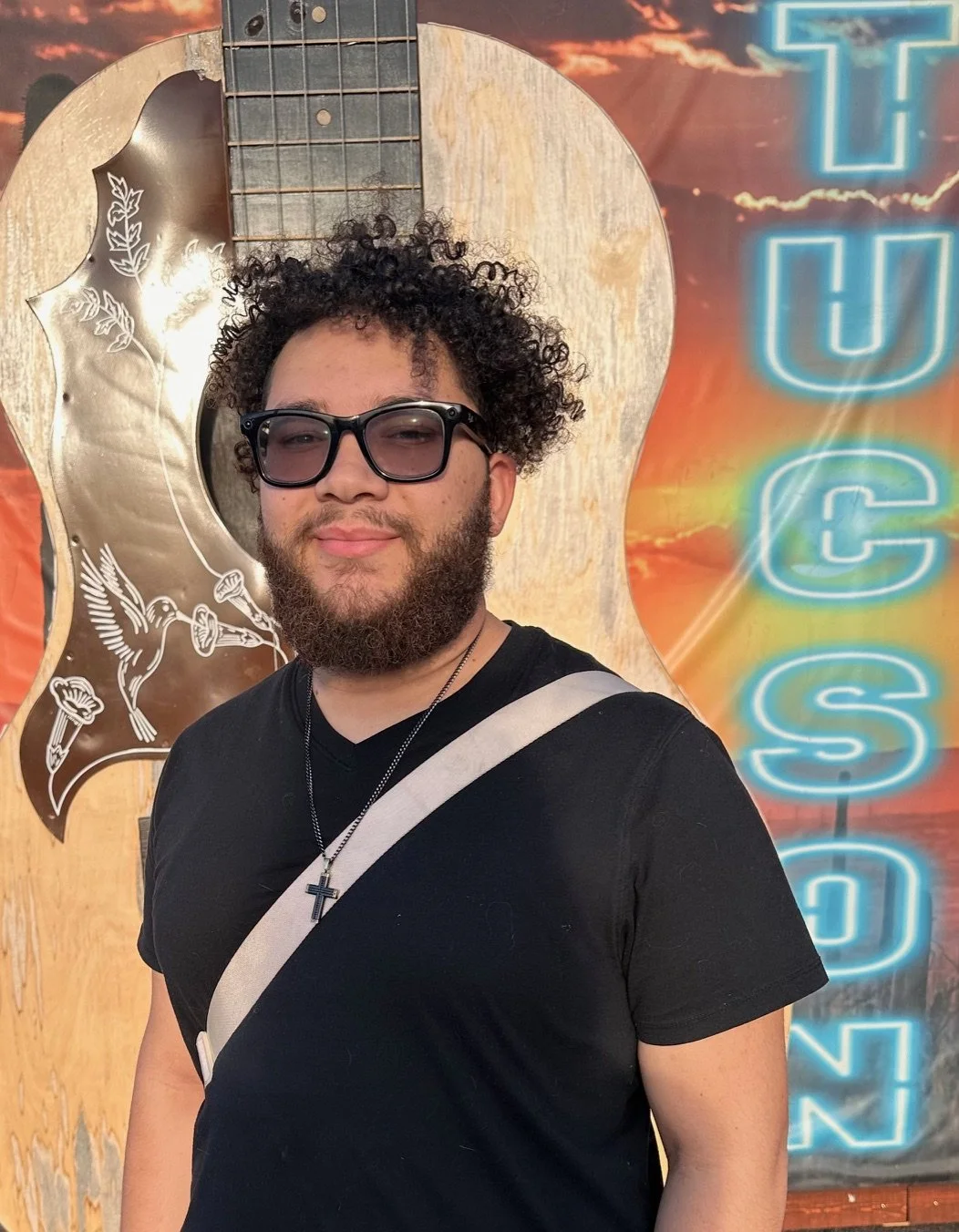 A young man with curly hair, sunglasses, and a beard, wearing a black shirt and cross necklace, standing in front of a decorative wooden guitar with floral designs and a neon sign that partially reads 'NASH'.