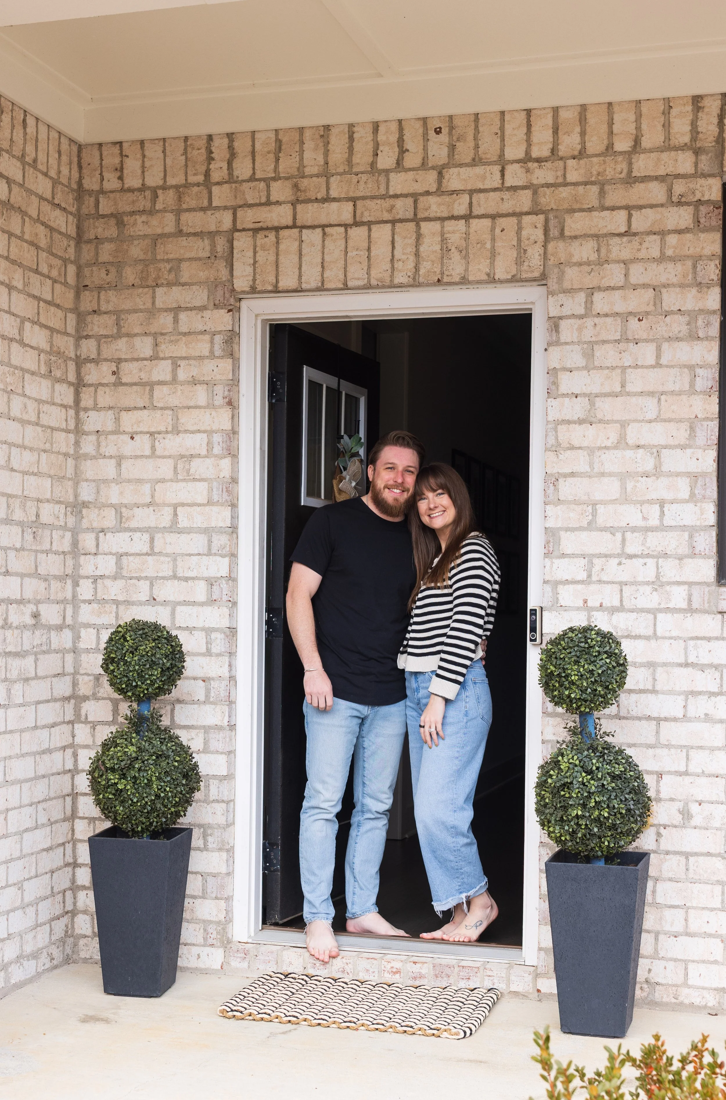 A smiling couple stands inside a doorway at the front of a house, with large potted topiary plants on either side of the entrance, on an outdoor porch.
