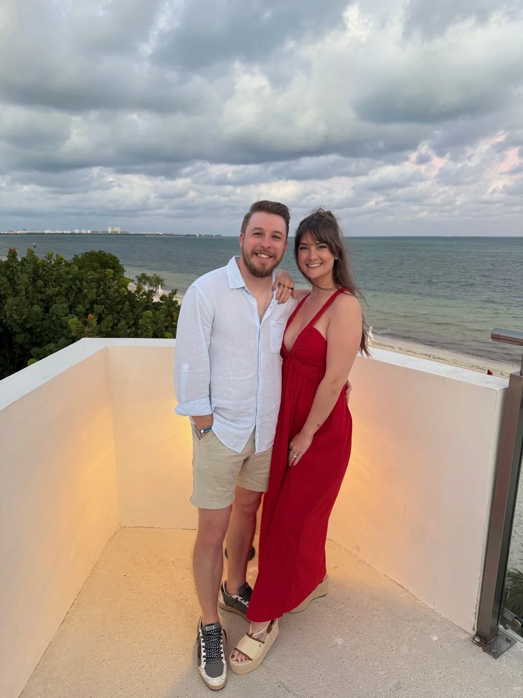 A smiling couple standing on a balcony with the ocean and cloudy sky in the background. The man is wearing a white long-sleeve shirt and beige shorts, and the woman is in a red dress. They are holding each other and smiling at the camera.