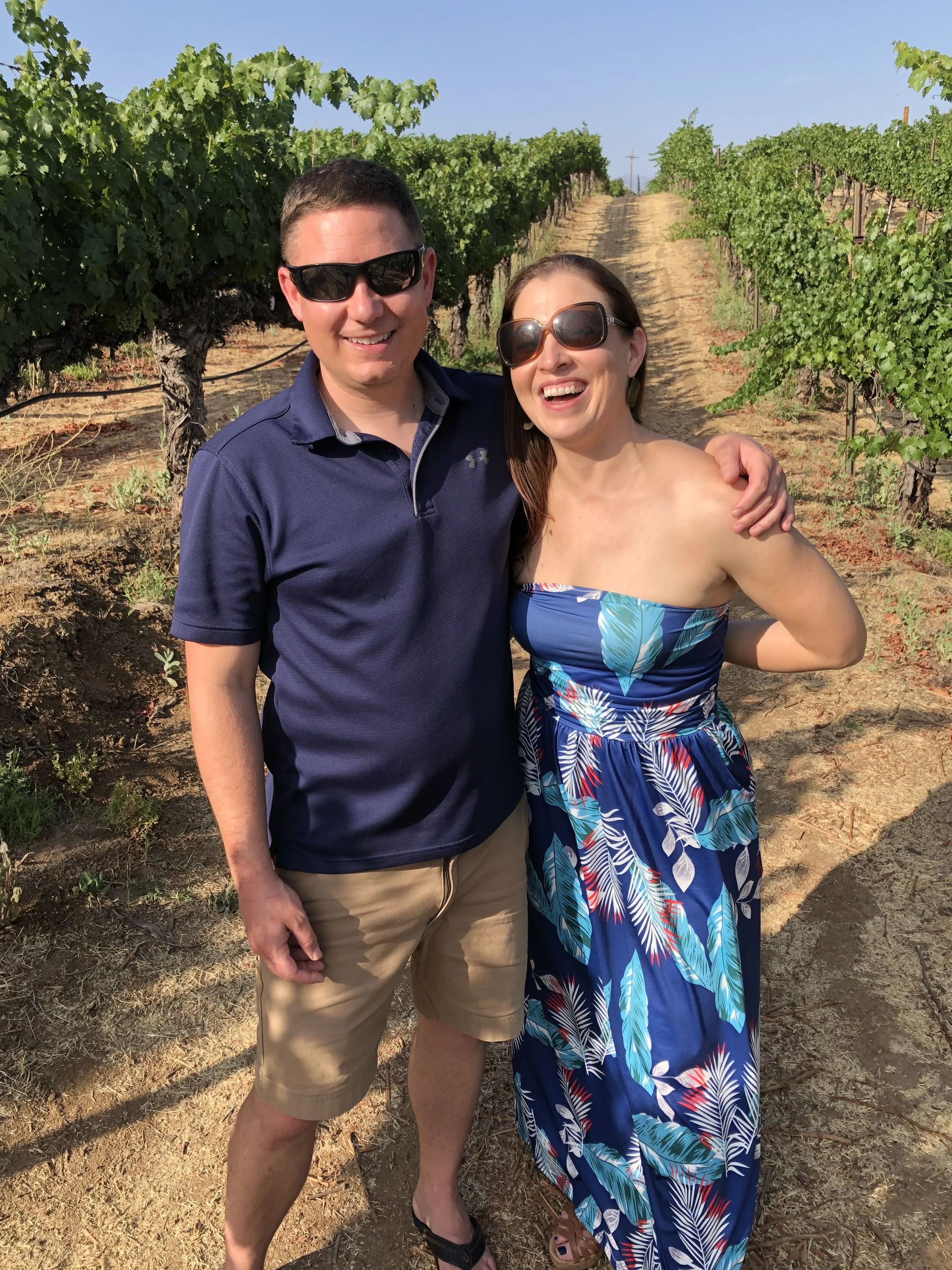 Two people smiling and embracing in a vineyard with green grapevines, dusty ground, and a clear blue sky in the background. considering adoption for her child and learning how to get pregnant help options to create an adoption plan.