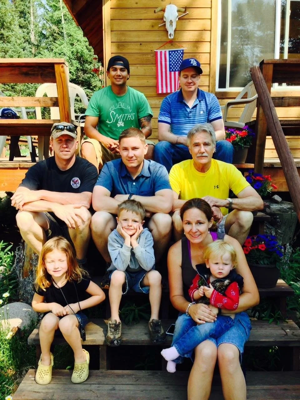 A family group photo on the steps of a wooden house with a carved animal skull and an American flag on the wall behind them. The group includes seven adults and two children, with various expressions and casual clothing. There are potted plants and flowers around them.