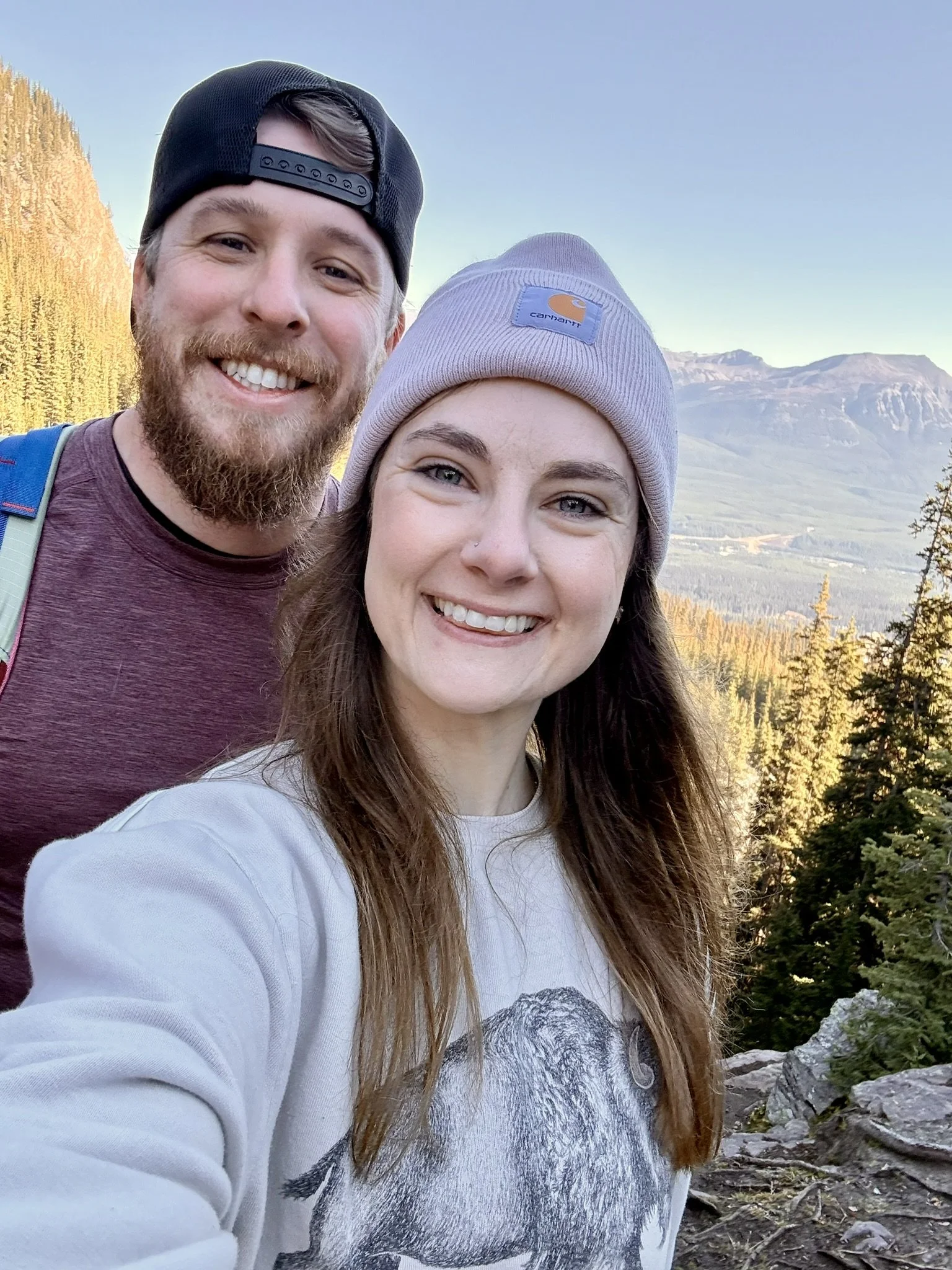 A smiling man and woman taking a selfie outdoors in a mountainous area during daytime with rocky terrain and trees in the background.