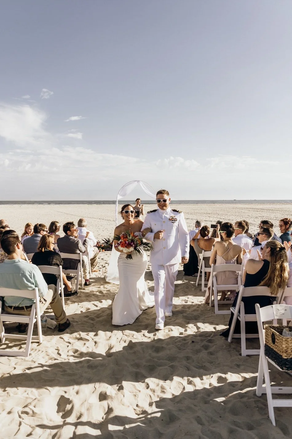 A beach wedding ceremony with a bride and groom walking down the aisle, surrounded by seated guests, on a sandy beach with the ocean and a partly cloudy sky in the background. considering adoption for her child and learning how to get pregnant help o