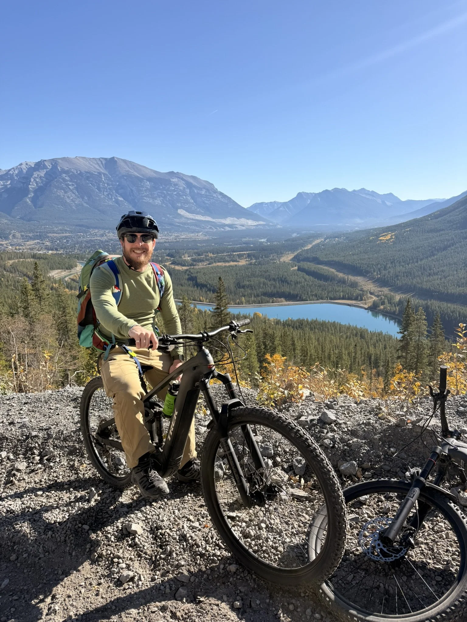 A man with a beard, sunglasses, and a helmet sitting on an electric mountain bike, smiling, with a scenic mountain landscape and a lake behind him.
