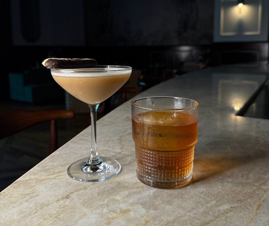 Two cocktails on a bar counter: a cocktail in a coupe glass with a dark chocolate garnish and a brownish layer, and a short glass with a warm amber-colored drink and ice cubes.
