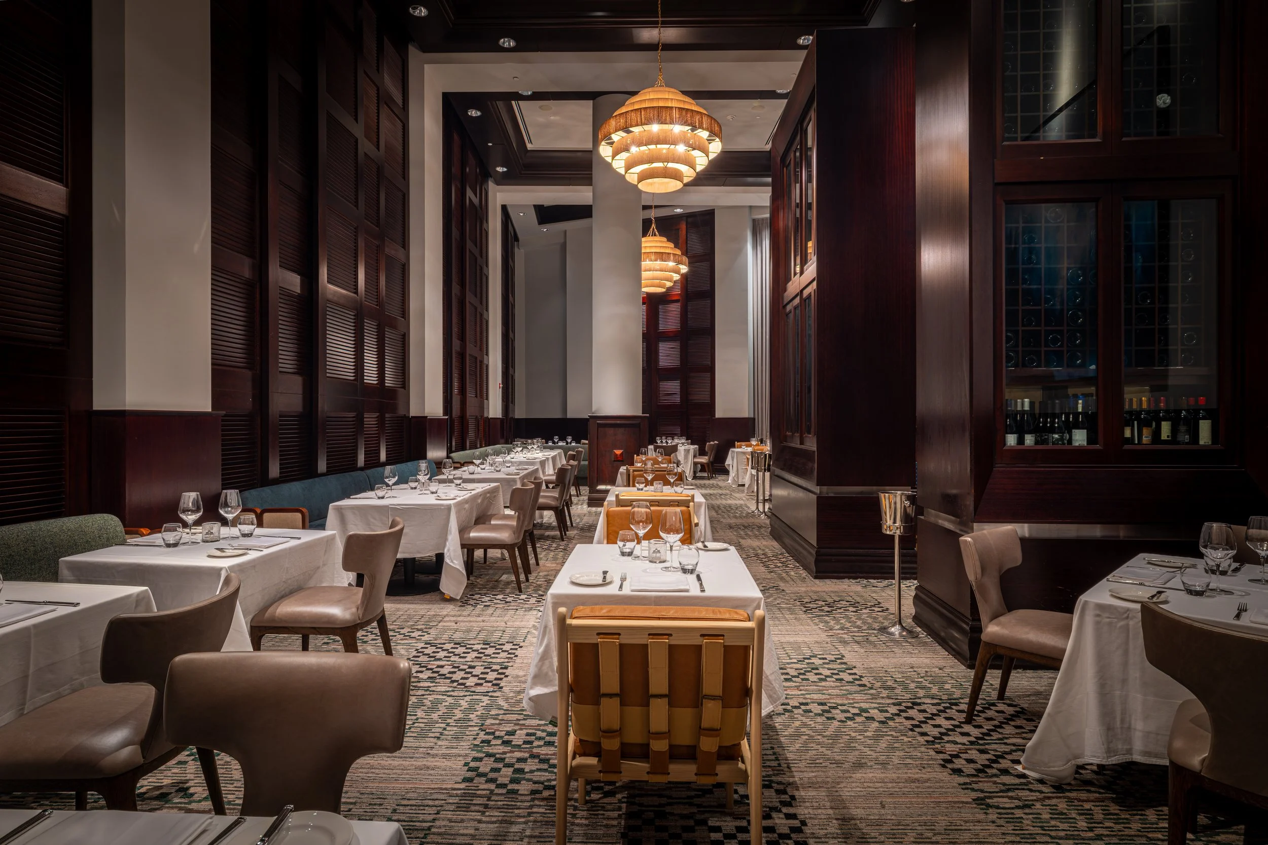 Elegant restaurant interior with white tablecloths, glasses, silverware, and various chairs, wooden paneling, and modern ceiling lights.