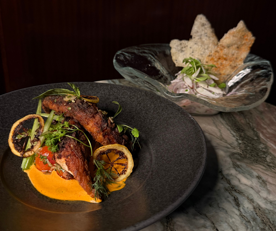 A gourmet dish featuring crispy caramelized fish fillets garnished with lemon slices, herbs, and vegetables, served with a side salad in a decorative glass bowl with sliced onions and crackers.