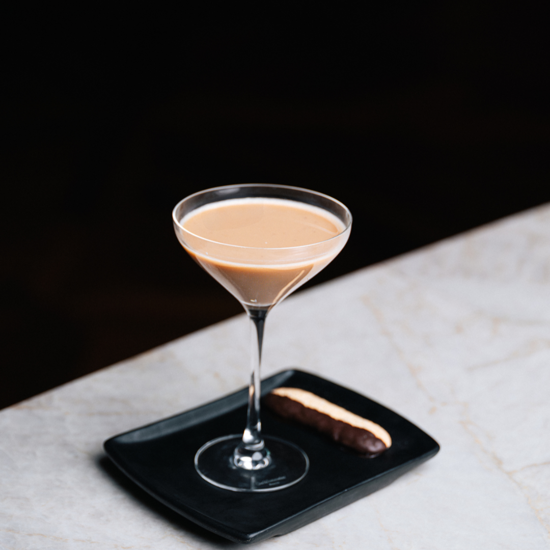 Martini glass filled with a light brown cocktail on a black plate, with a chocolate-coated biscuit on the side, all on a white marble surface.