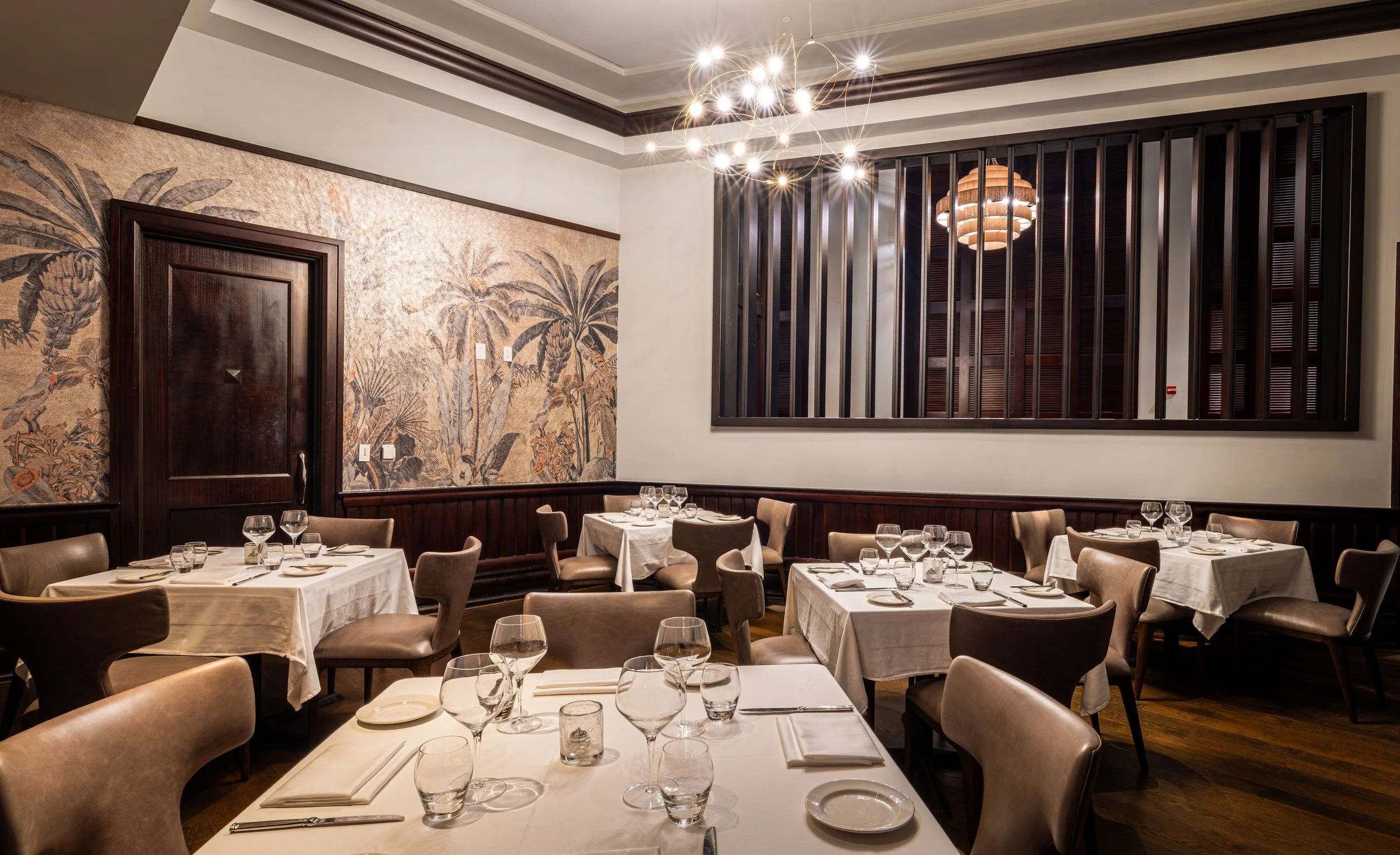 Elegant restaurant dining room with tables set with white tablecloths, glassware, and silverware. Dark wood accents, tropical mural on the wall, modern chandelier, and decorative wooden panel on the wall.