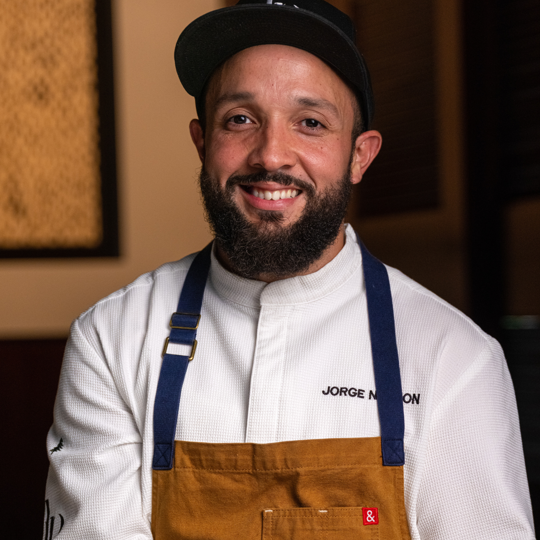 Portrait of a smiling male chef with a beard, wearing a black cap, white chef's coat with his name "JORGE N" embroidered, and a brown apron with blue straps, in a restaurant setting.