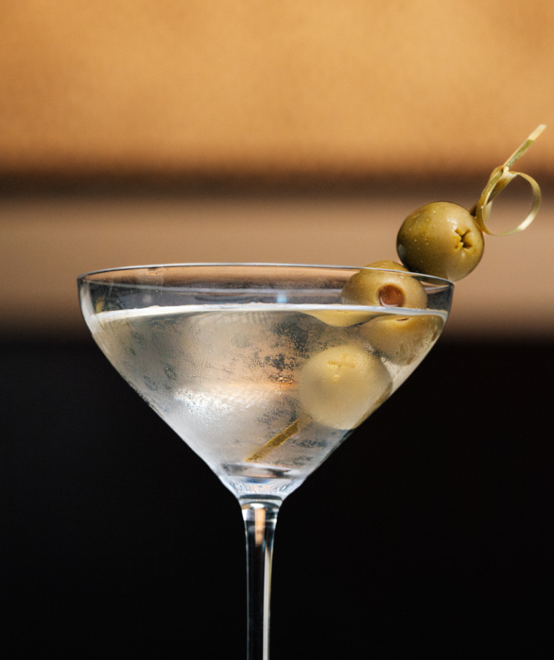 A martini glass with three green olives on a pick, served in a clear cocktail glass