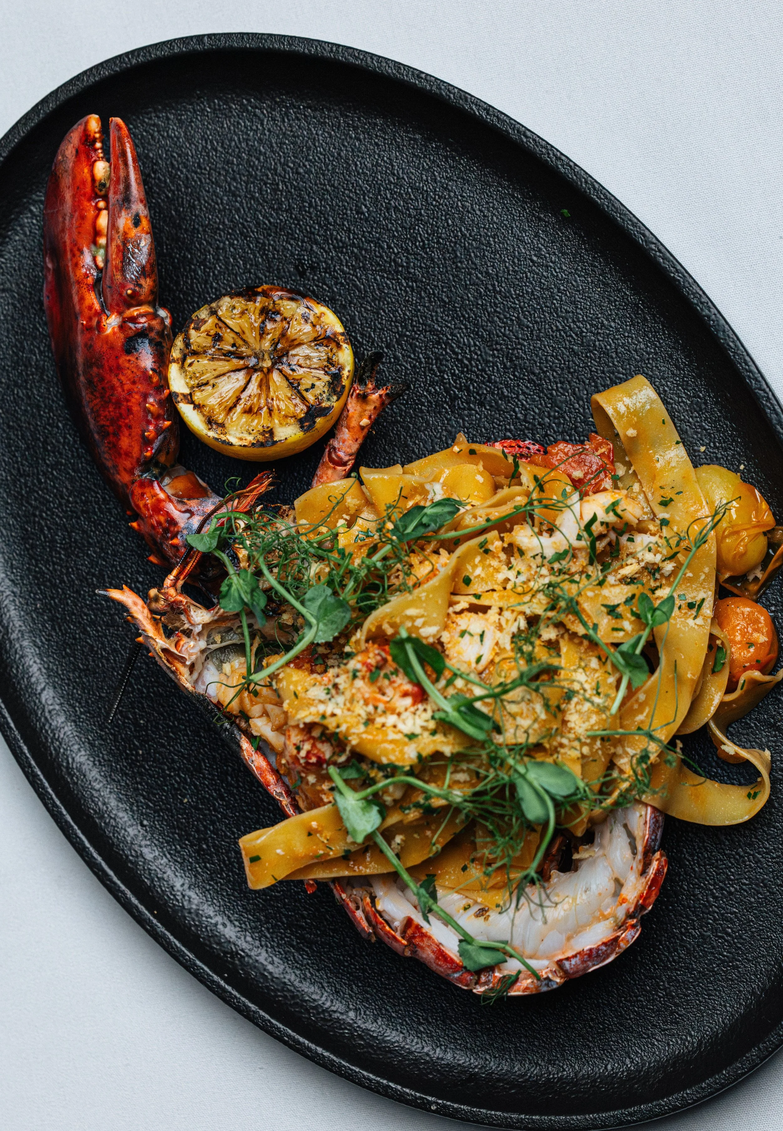 Lobster tail with grilled lemon half and pasta with herbs on a black plate.