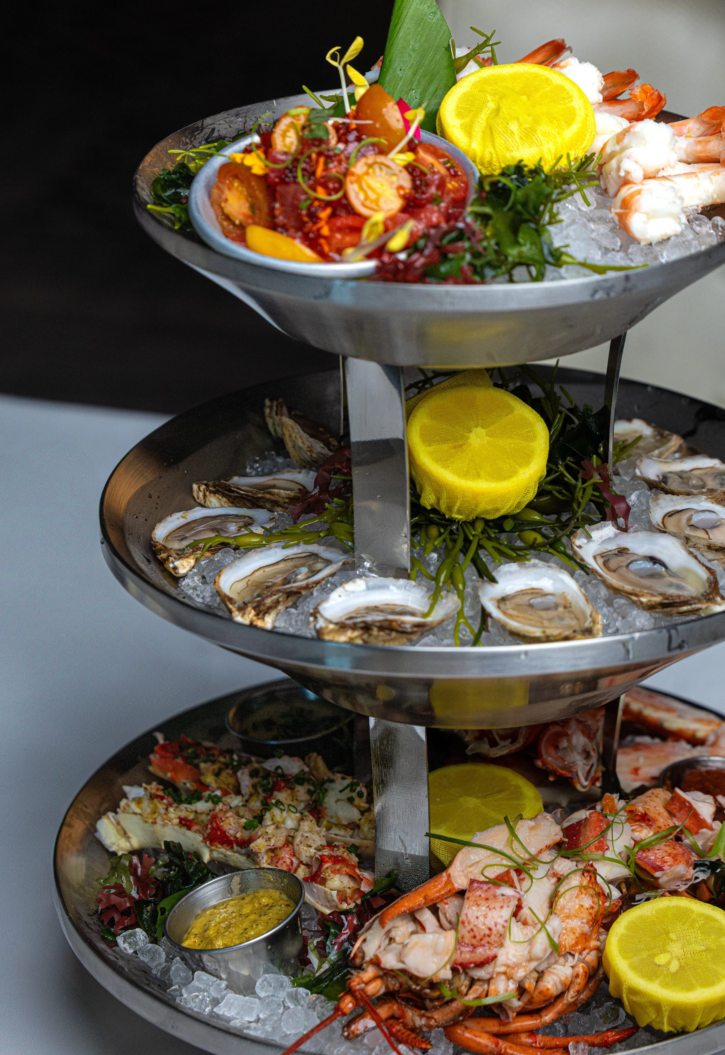 A multi-tiered seafood platter featuring oysters, shrimp, lobster, and crab, garnished with lemon slices and greens.