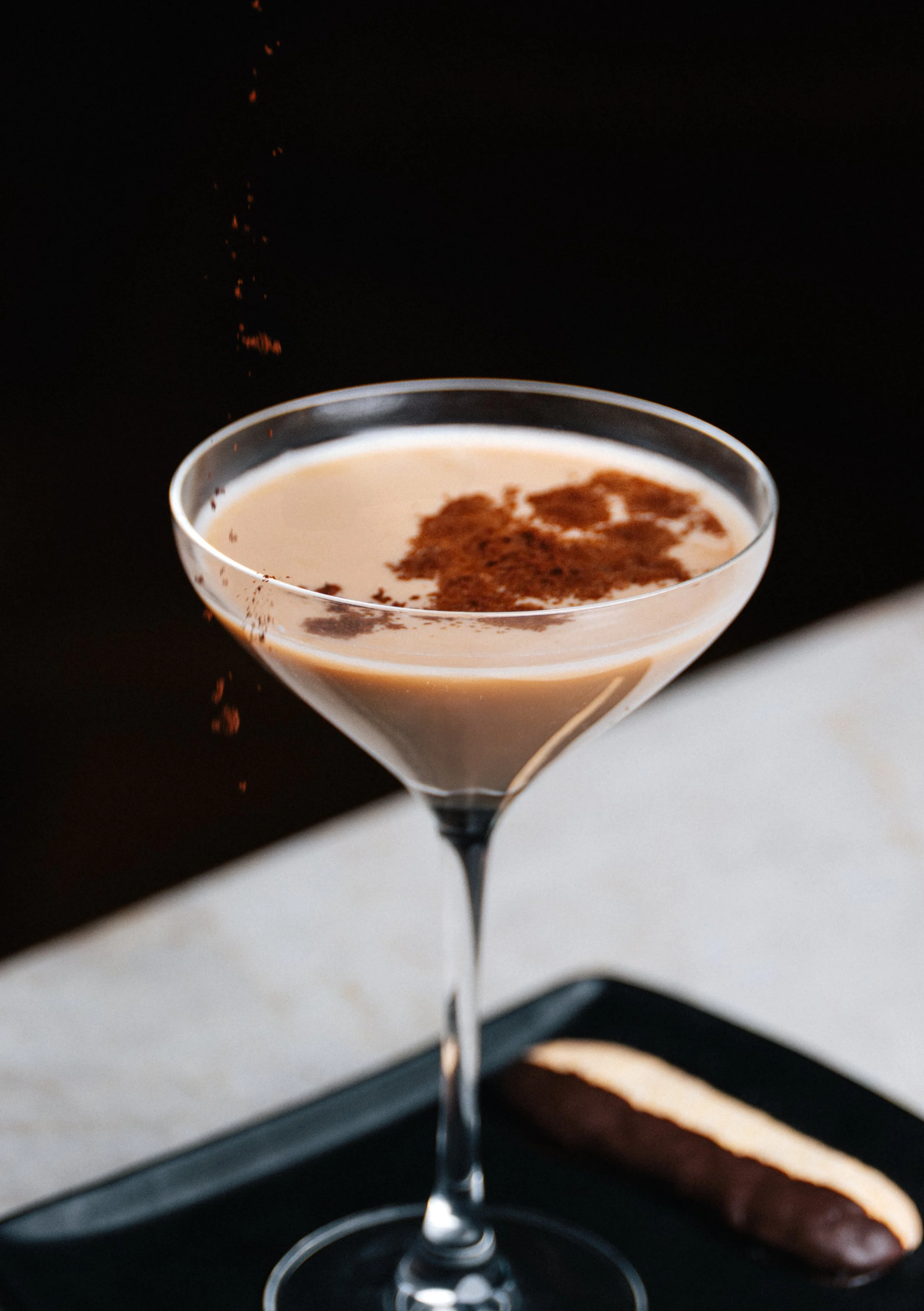 A cocktail glass filled with a creamy brown beverage, topped with a dusting of cocoa powder, sitting on a black tray with a chocolate-dipped biscuit beside it.