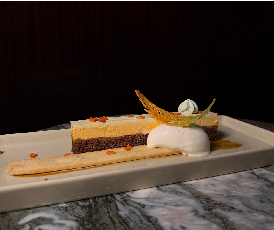 A slice of layered cheesecake on a white rectangular plate, garnished with a decorative leaf, whipped cream, and a drizzle of caramel, with small orange pieces garnish.