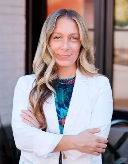 A woman with long blonde hair wearing a white blazer over a colorful top, standing outside with her arms crossed.