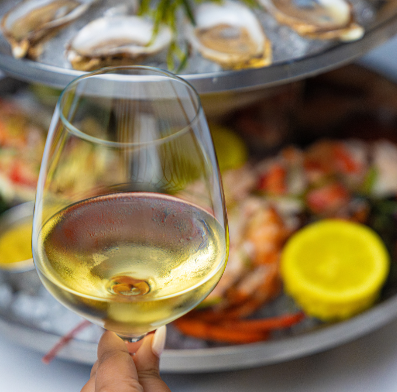 Close-up of a hand holding a glass of white wine with a seafood platter and lemon in the background.