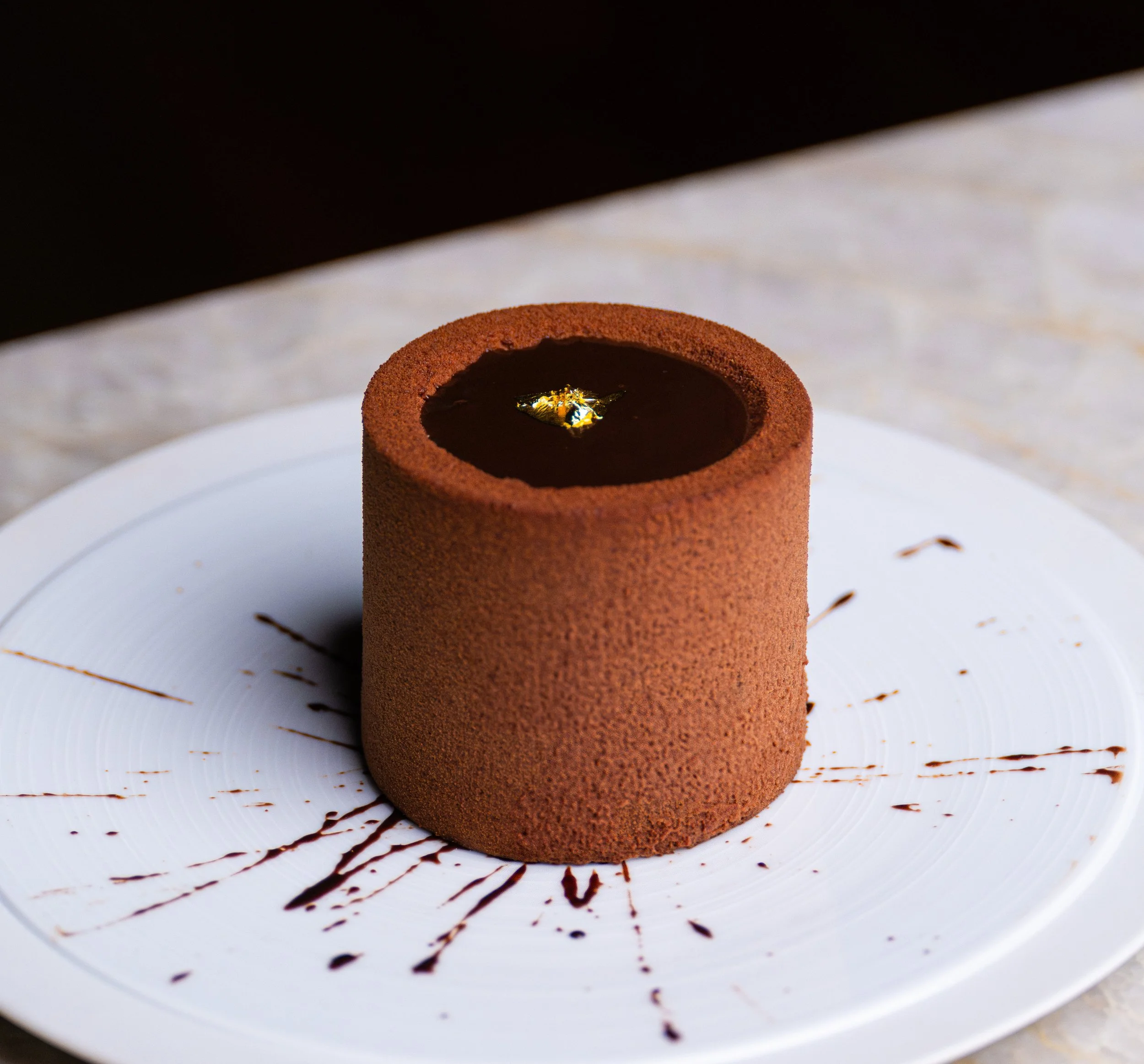 Chocolate mousse cake with gold leaf decoration on top, served on a white plate decorated with chocolate drizzle.