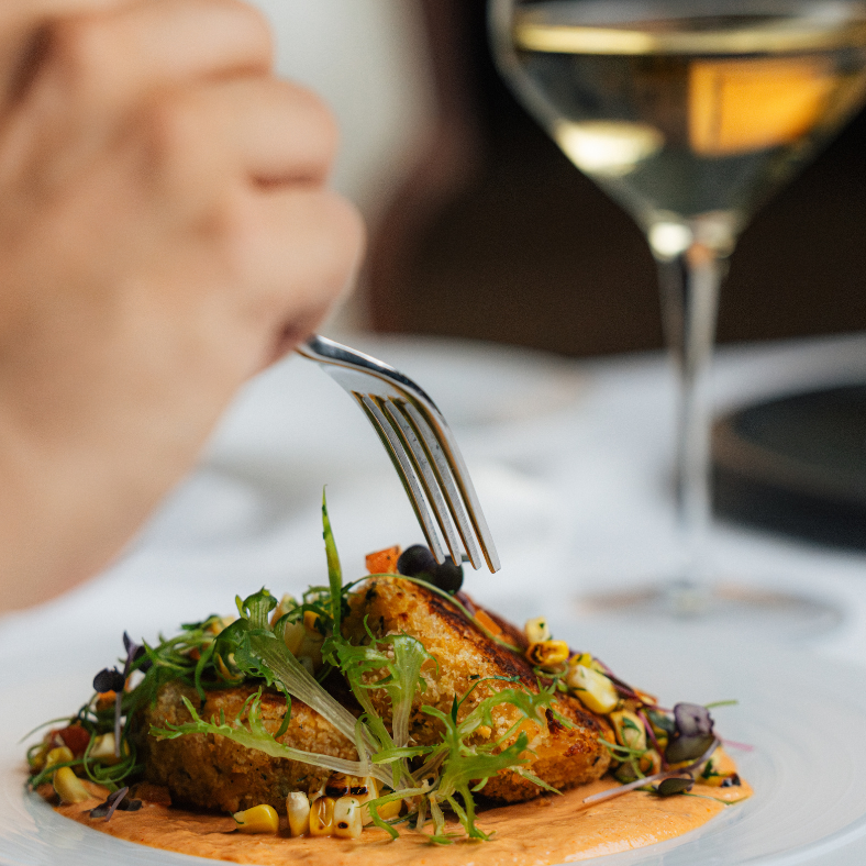 Person eating a plated dish with grilled fish, greens, and vegetables, accompanied by a glass of white wine.