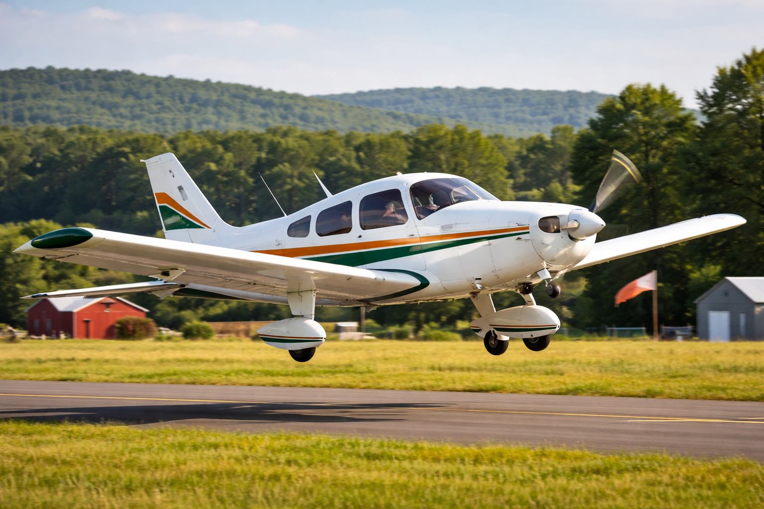 Small white airplane with green and orange stripes landing on a runway with green fields and trees in the background.