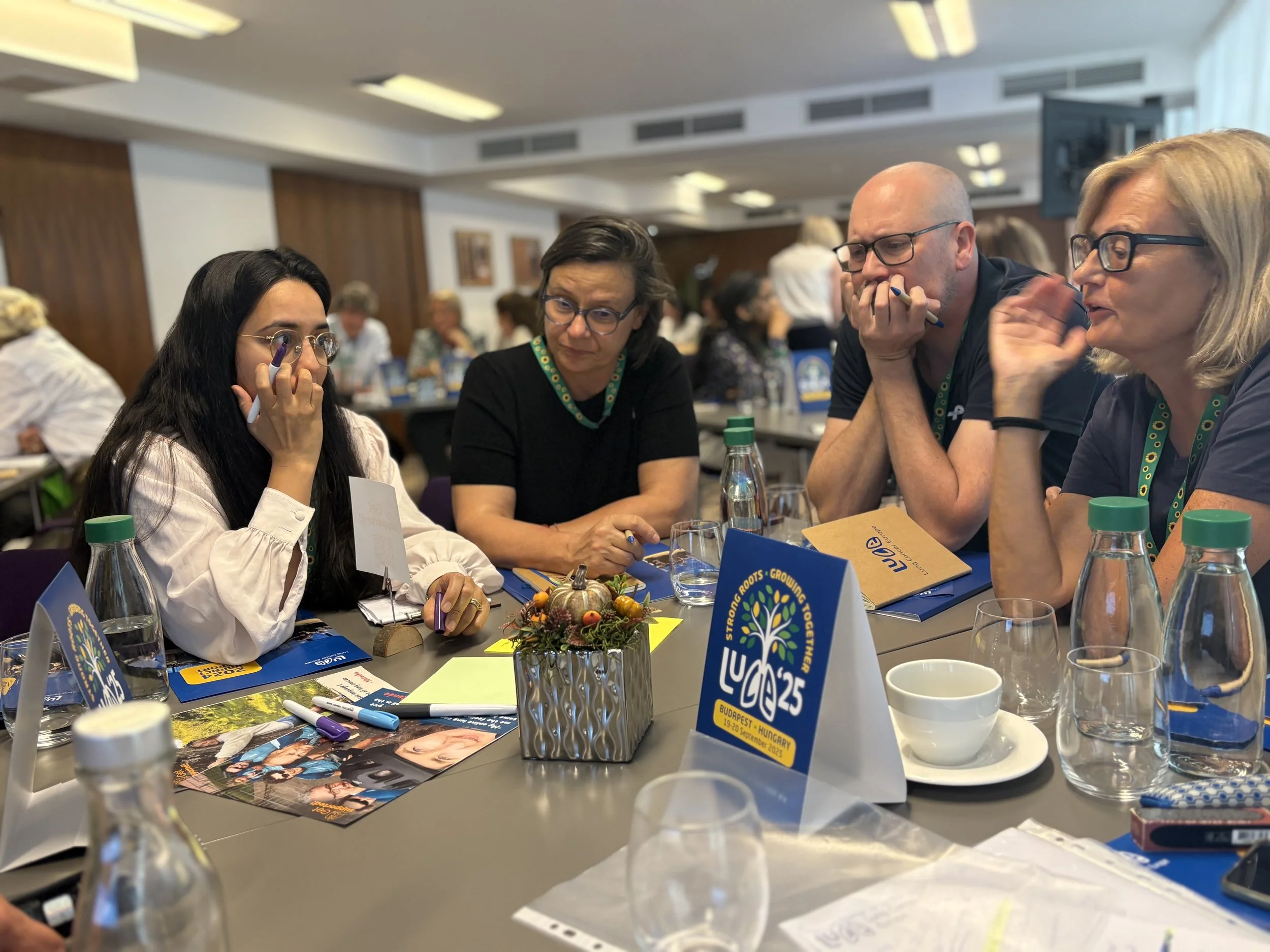 Four people sitting at a conference table engaging in discussion, with conference materials and water bottles on the table, and a decorative centerpiece with flowers and pumpkins.