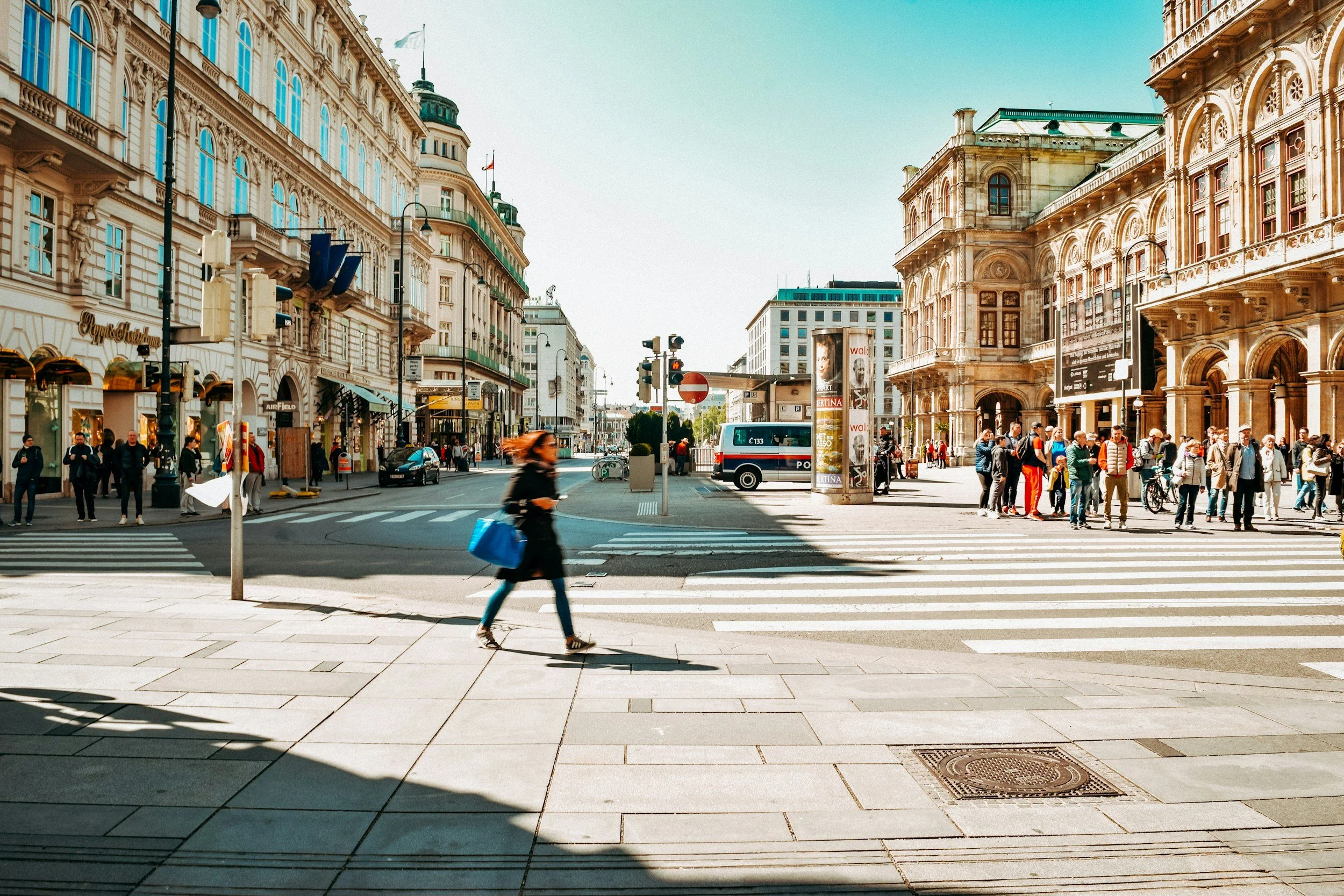 Crowded city street scene with historic buildings, pedestrians crossing crosswalk, and a police van parked on the side.