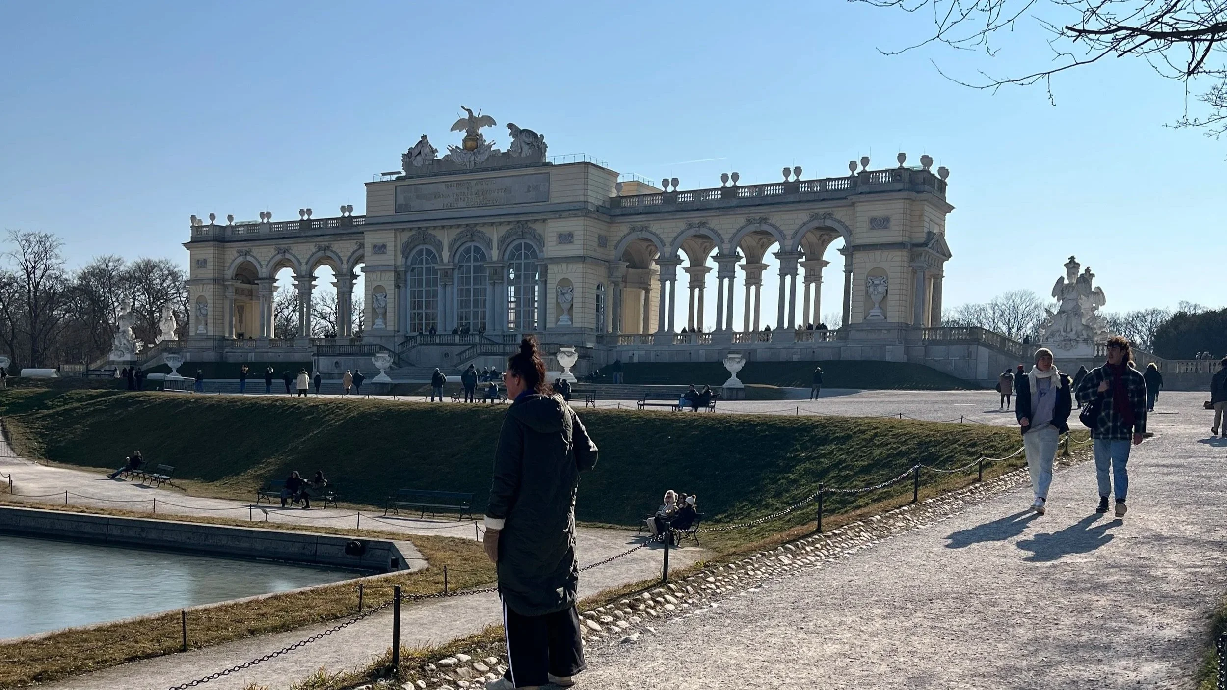 People walking on a gravel path in front of a large, ornate palace with arches, columns, and sculptures, on a clear sunny day.
