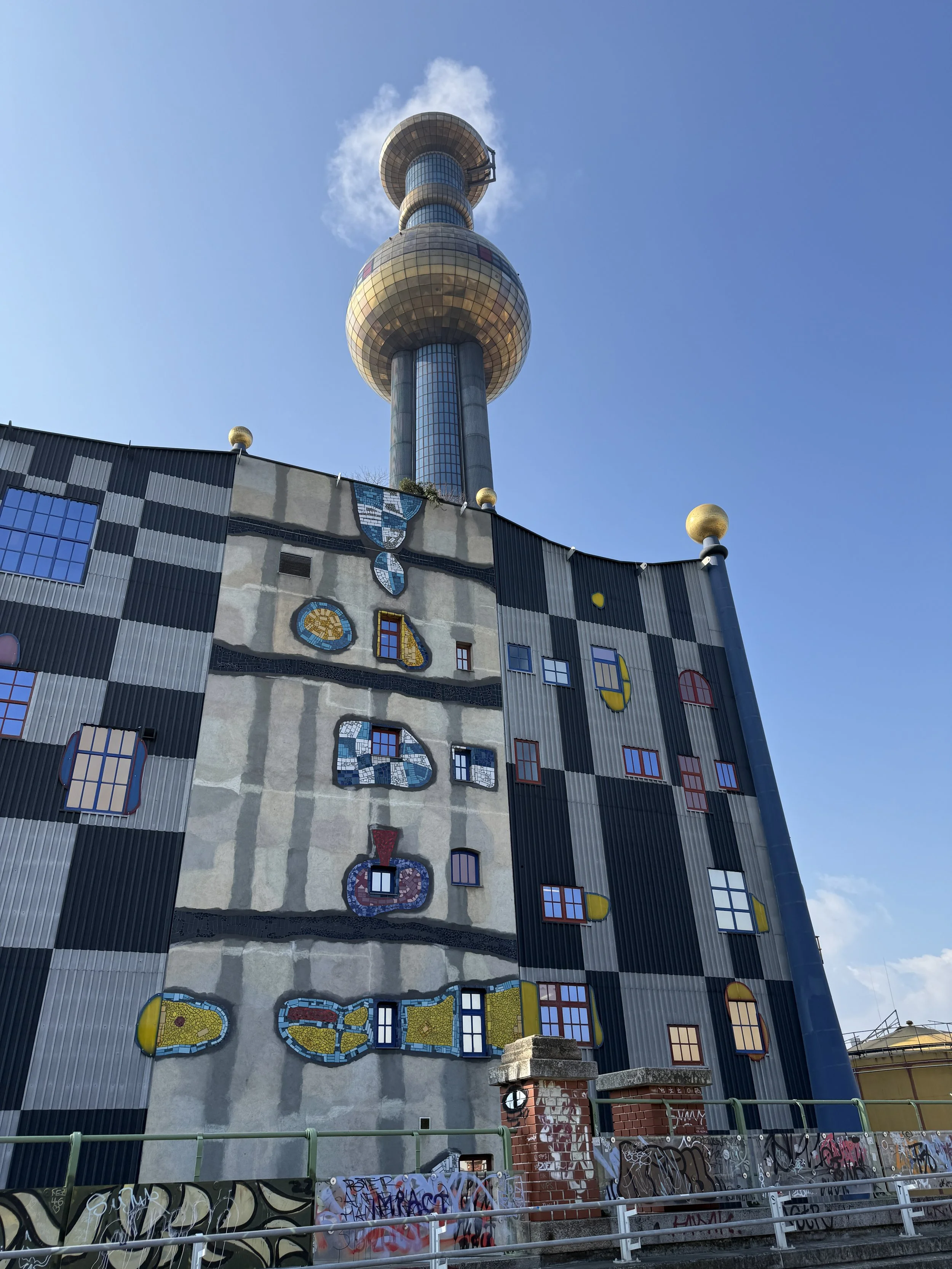 A colorful, eclectic building with a large tower resembling a radio or TV tower on top, featuring a spherical section and a smaller dome. The building's facade has abstract, whimsical patterns, windows, and decorative elements, with some graffiti on the lower part.