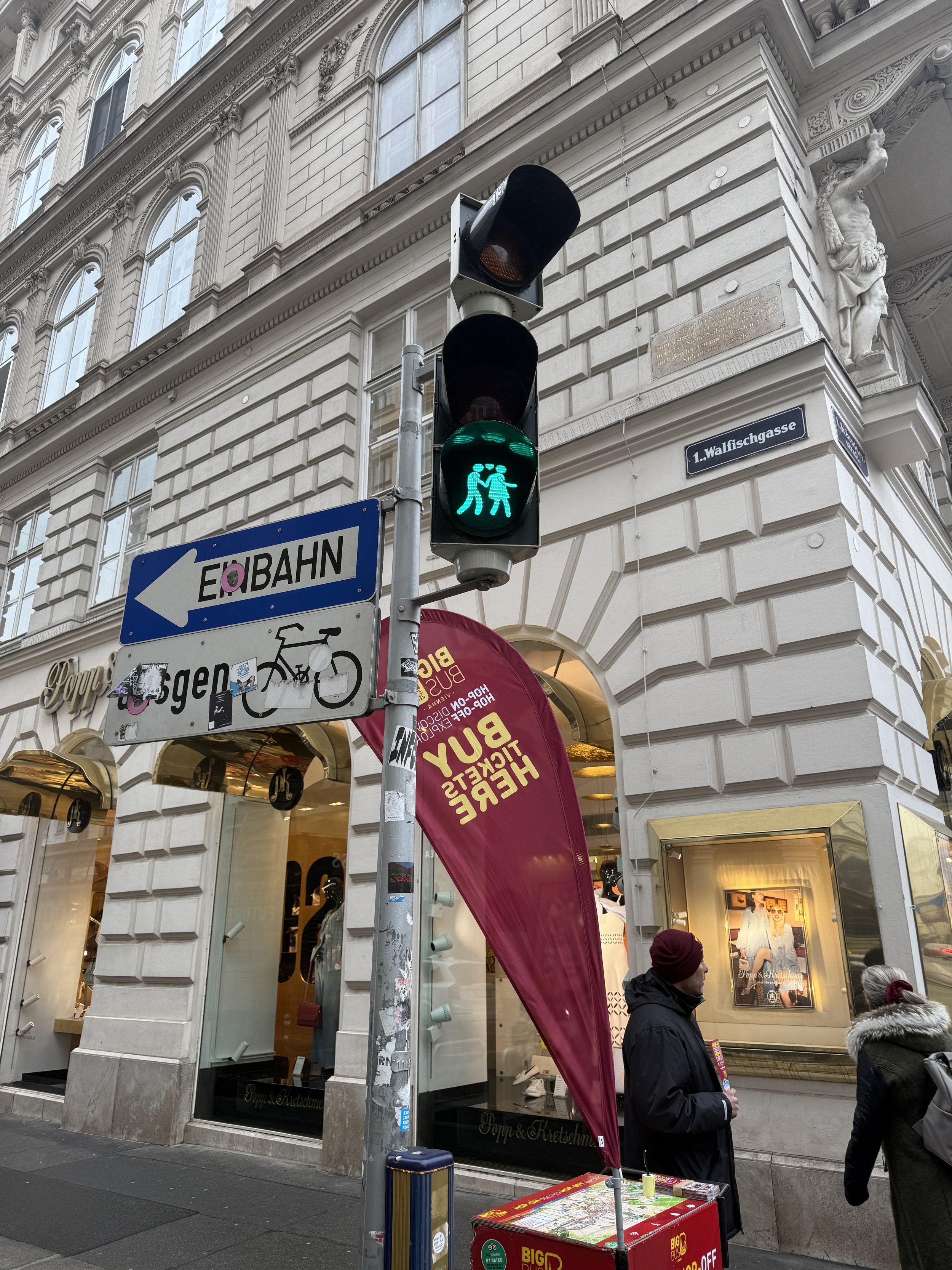Street scene in front of a jewelry store with pedestrians, a traffic light from Vienna's famous inclusive initiative showing green for pedestrians, signage, and a billboard. The building features ornate architecture and a sculpture on the corner.