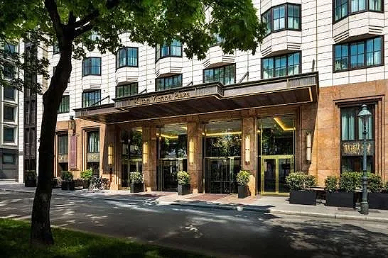 Entrance of a modern building with large glass doors and windows, surrounded by potted plants, with a tree and street in the foreground.