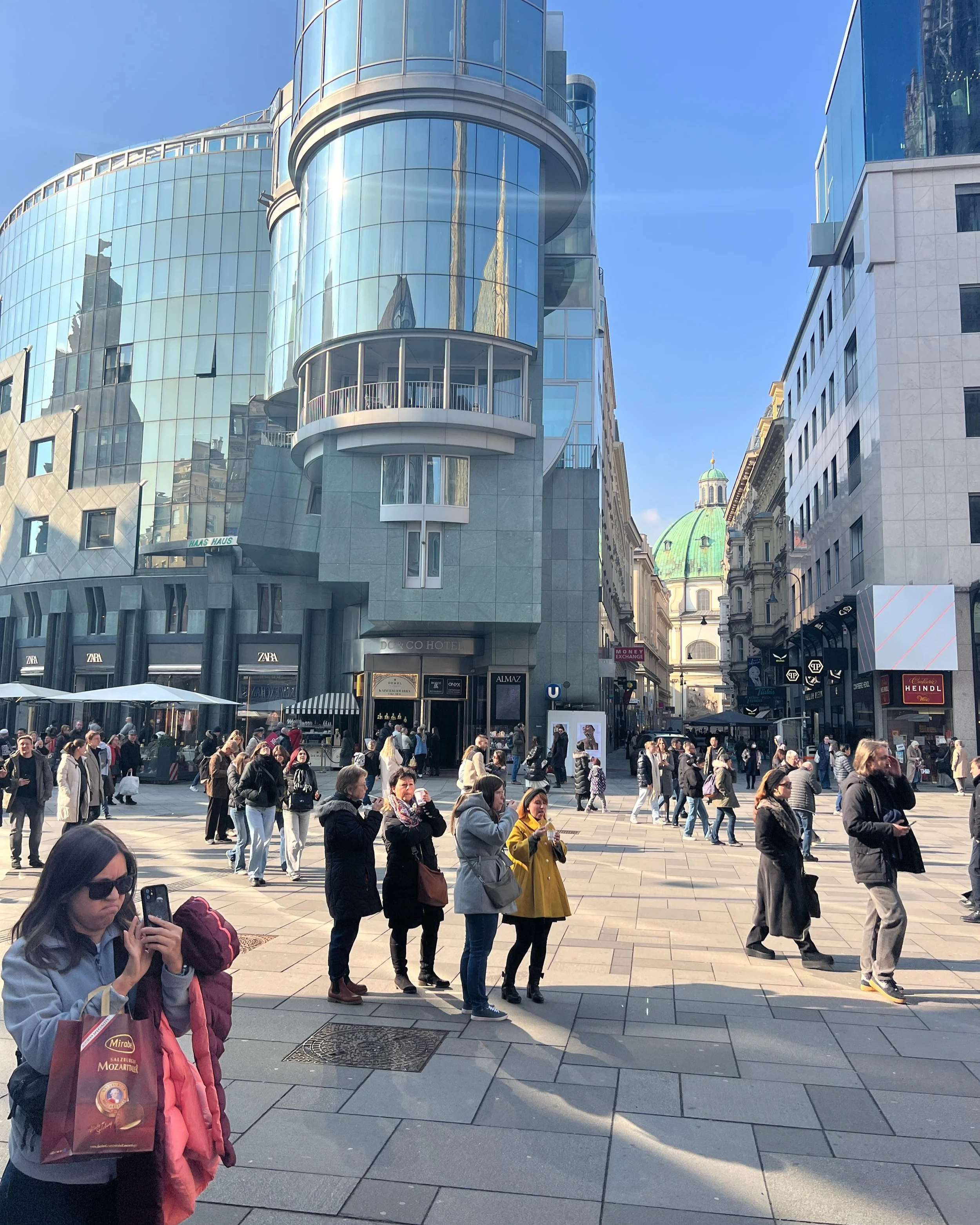 Crowd of people walking and standing in a busy urban square with modern glass buildings and a church with a green dome in the background.