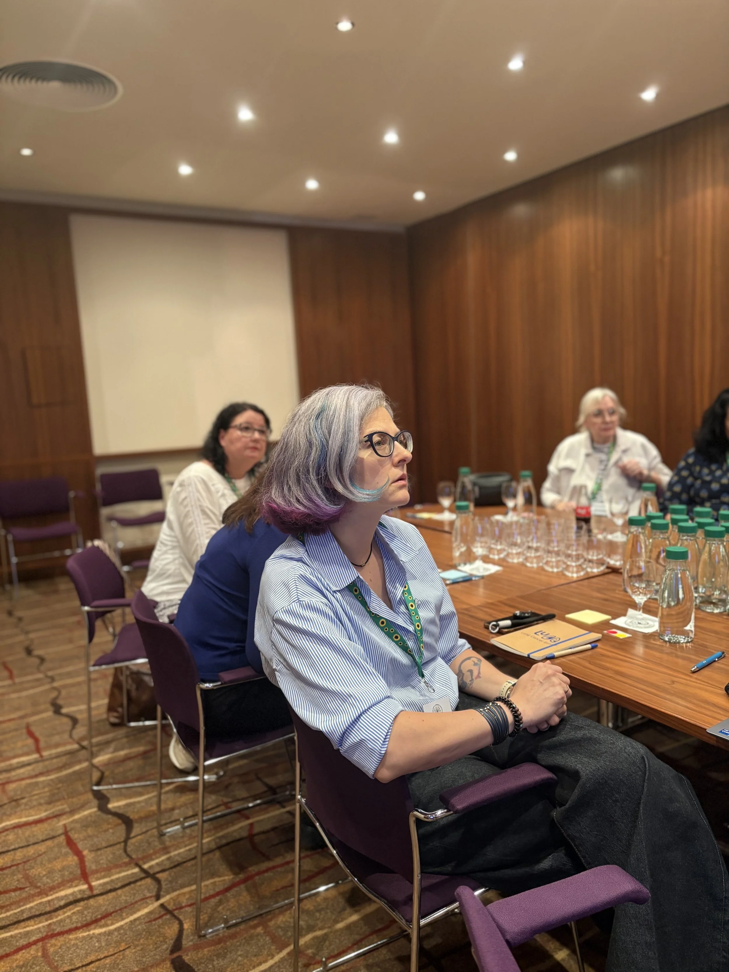 A group of women sitting around a conference table in a wood-paneled room, attentively listening to a speaker, with water bottles, glasses, and notebooks on the table.