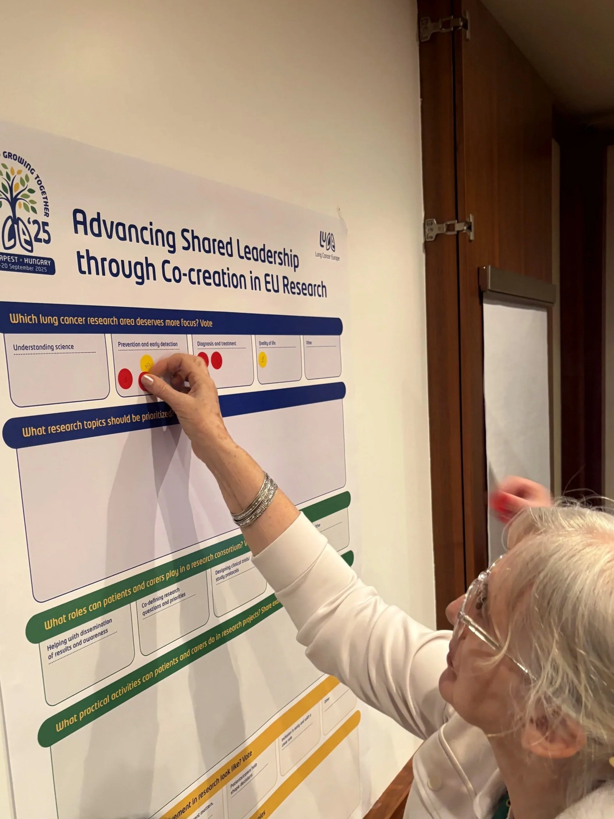 An elderly woman with glasses is placing a red sticky note on a research poster about EU research collaboration in lung cancer.