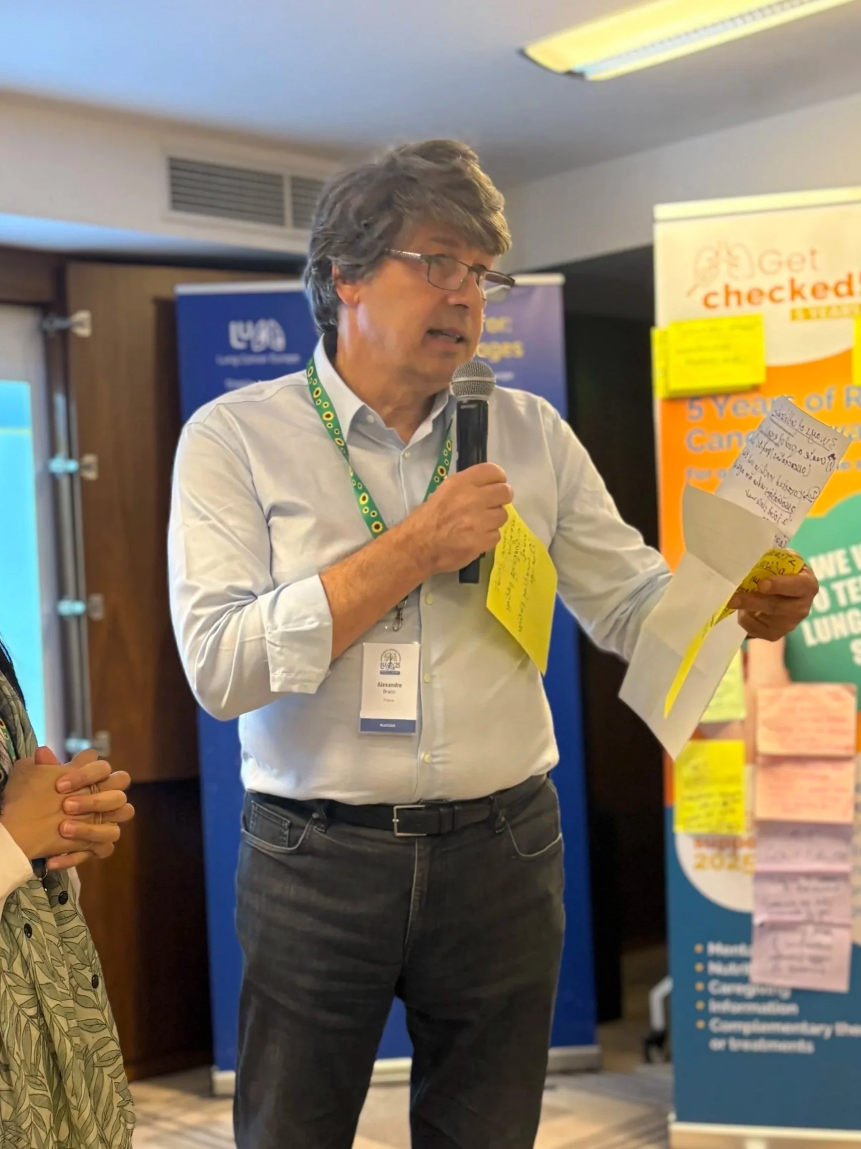 A man with gray hair and glasses is speaking into a microphone, holding a note and wearing a name badge. Behind him are banners with information, indicating it is an indoor event or conference.