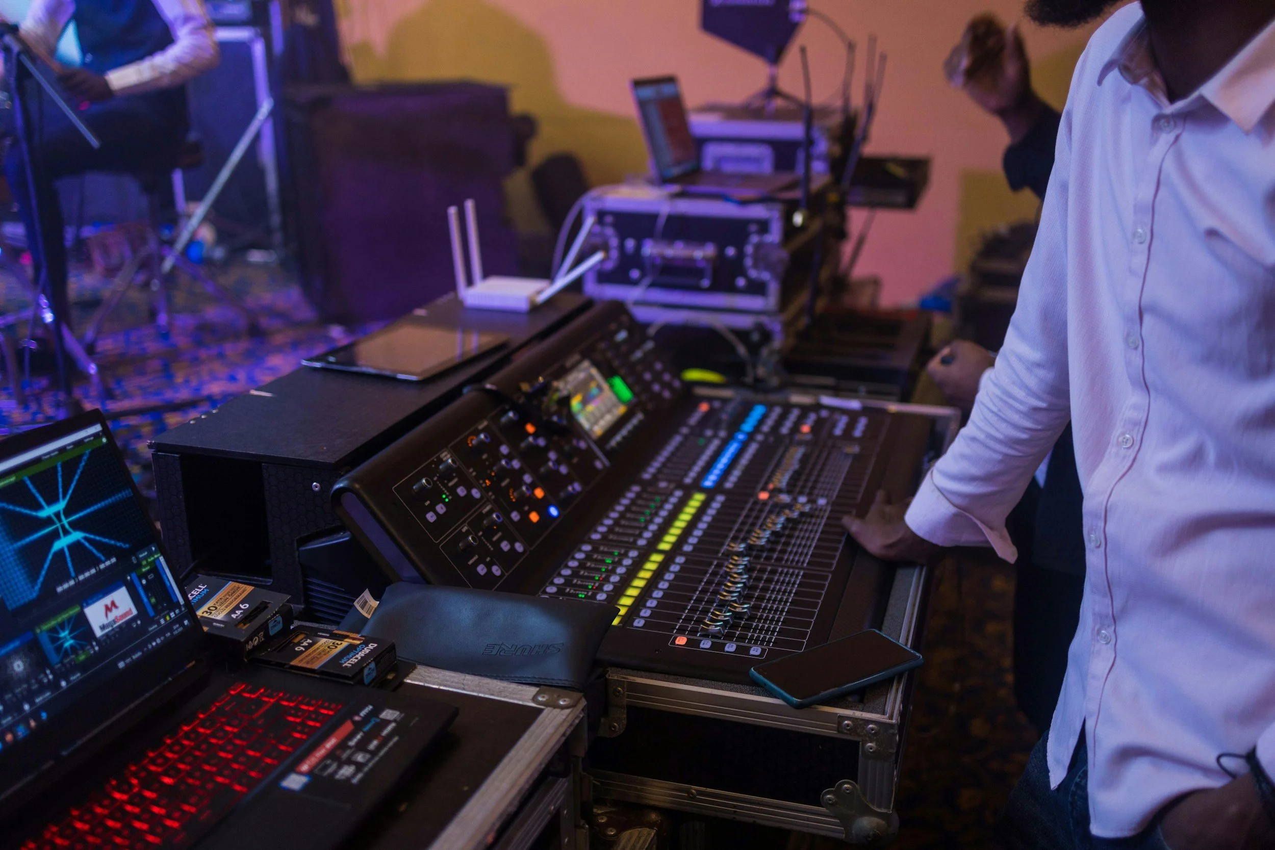 A person standing at a sound mixing console in a music studio or live event setup, with other audio equipment and a laptop nearby.