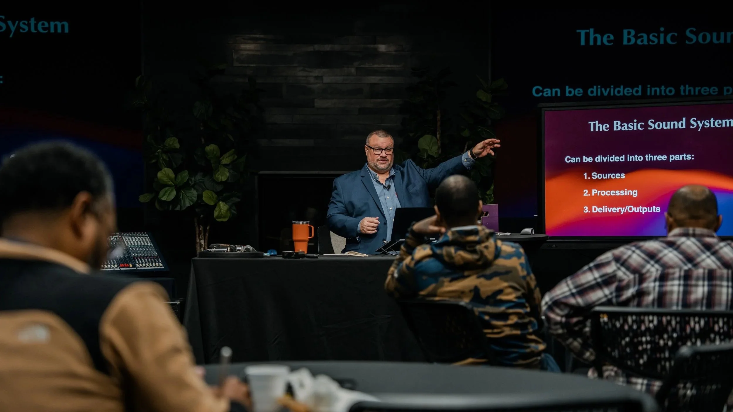 Ike giving a lecture in front of an audience, pointing towards a screen displaying a presentation on the basic sound system, while attendees watch and take notes.