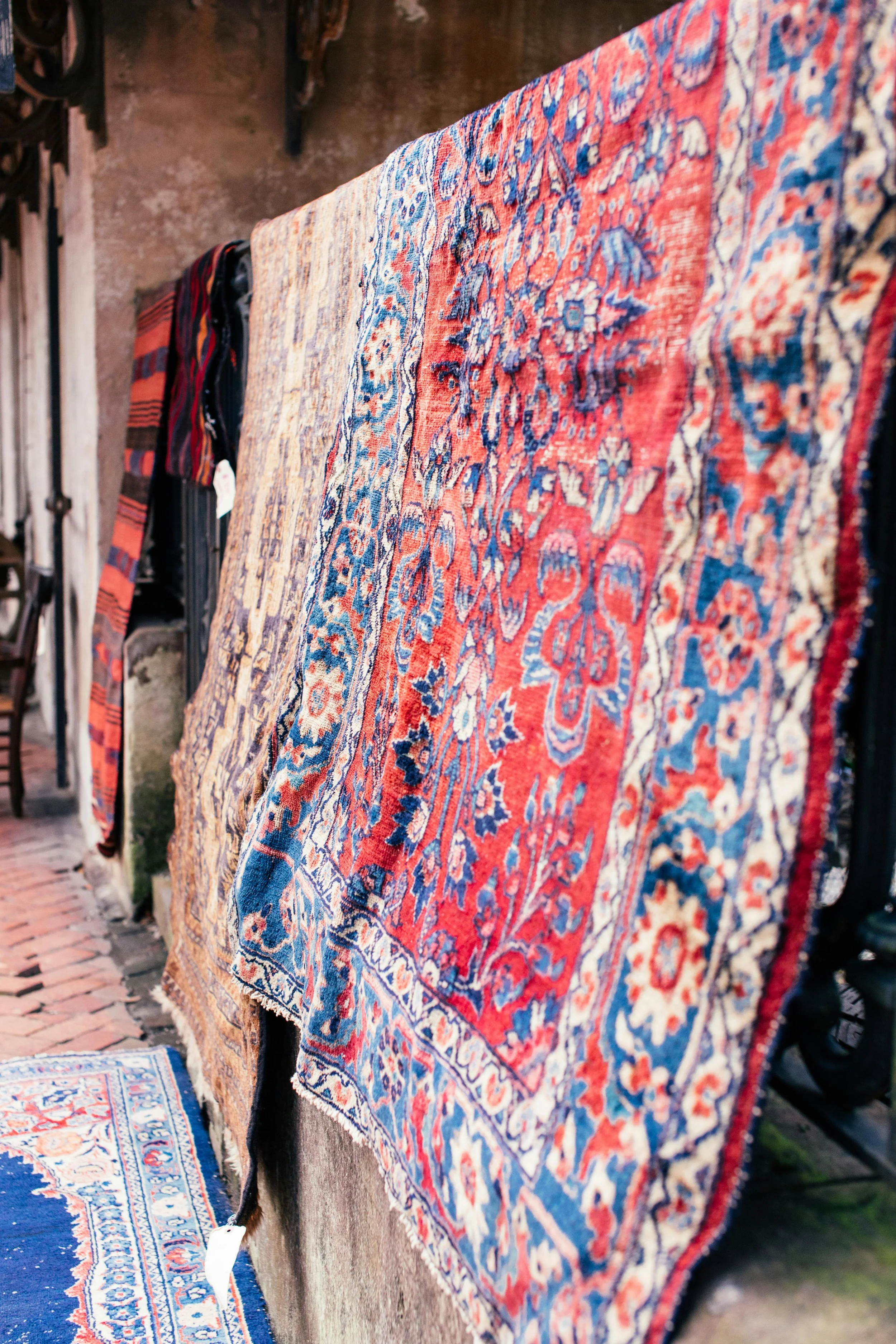 A display of vintage Persian rugs hanging on a wall outside an antique or thrift store, featuring colorful intricate patterns in red, blue, and beige.