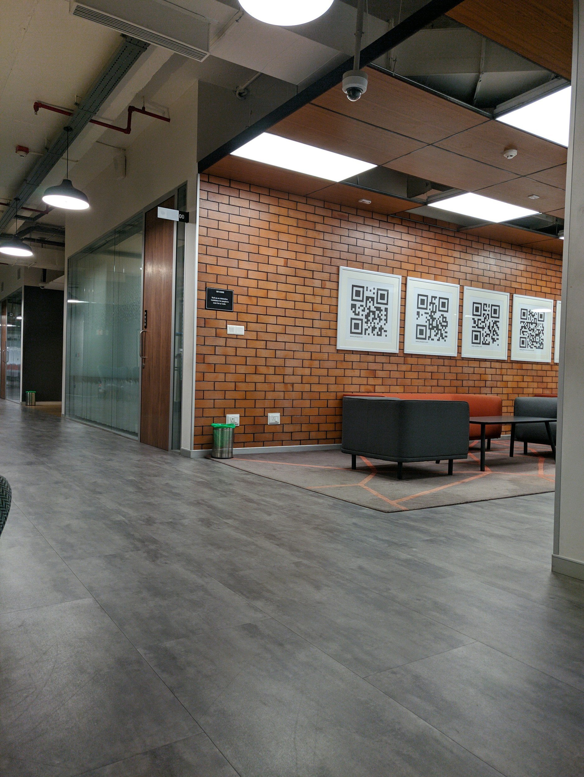 Modern office lounge area with black and orange chairs, a small table, a brick wall with framed QR code posters, and wooden ceiling panels.