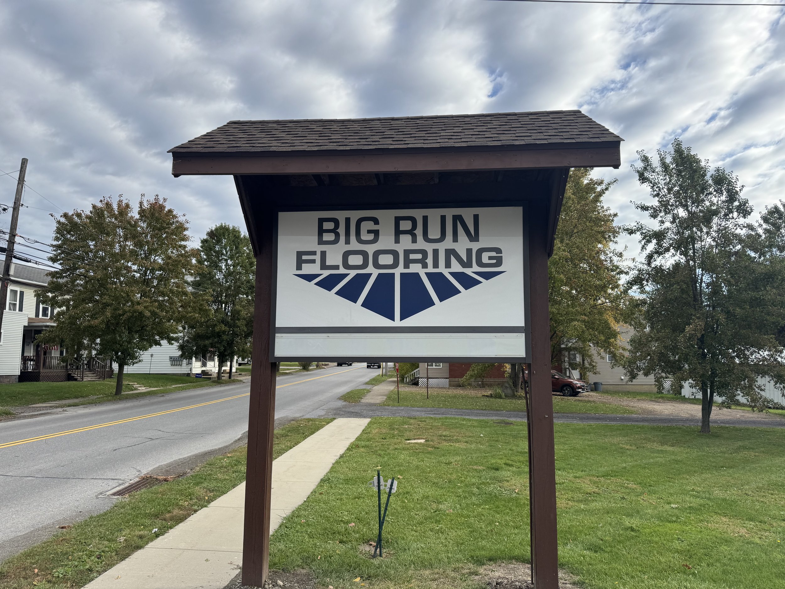 Sign for Big Run Flooring with a stylized blue and black logo, located on a sidewalk in a suburban neighborhood with trees and houses in the background.