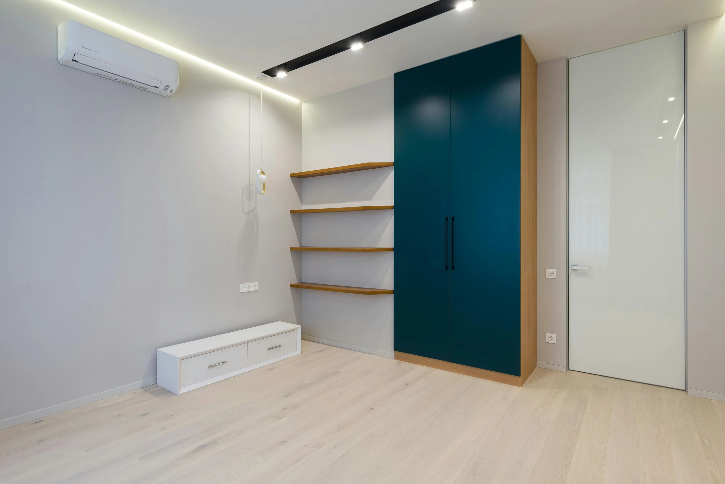 Empty modern room with white walls, light wood flooring, a white air conditioning unit, black ceiling lights, three wooden shelves attached to the wall, a large blue wardrobe with black handles, a white door, and a small white cabinet with drawers.