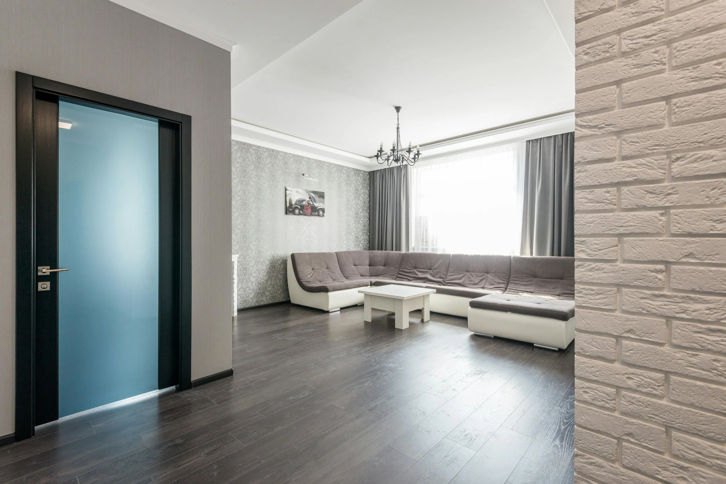 Modern living room with gray L-shaped sectional sofa, white coffee table, patterned accent wall, chandelier, large window with curtains, and a blue door.