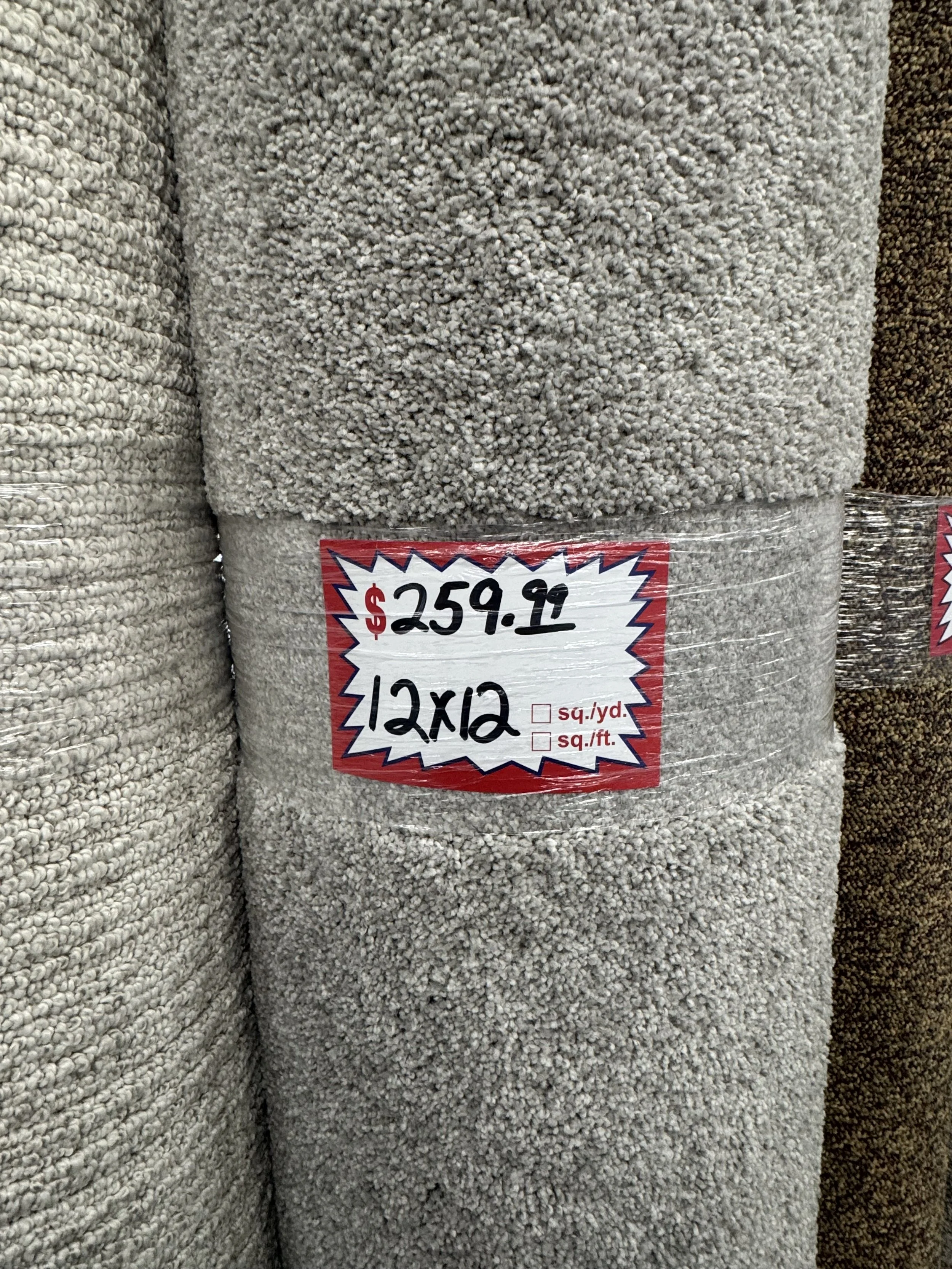 A beige textured carpet roll with a red and white price tag showing $259.99, size 12x12, and checkboxes for square yard and square feet.