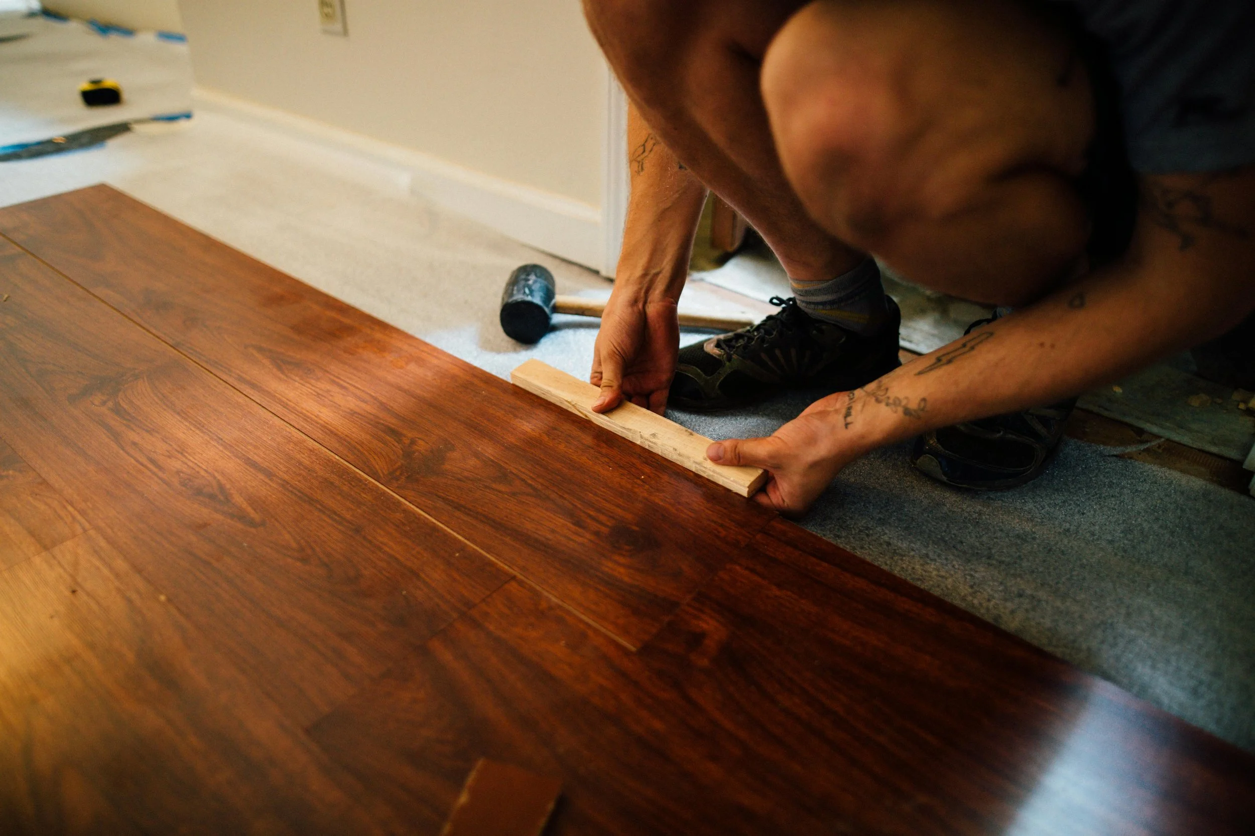A person is installing a wooden floor, using a piece of wood and a hammer on top of a light-colored carpeted room.